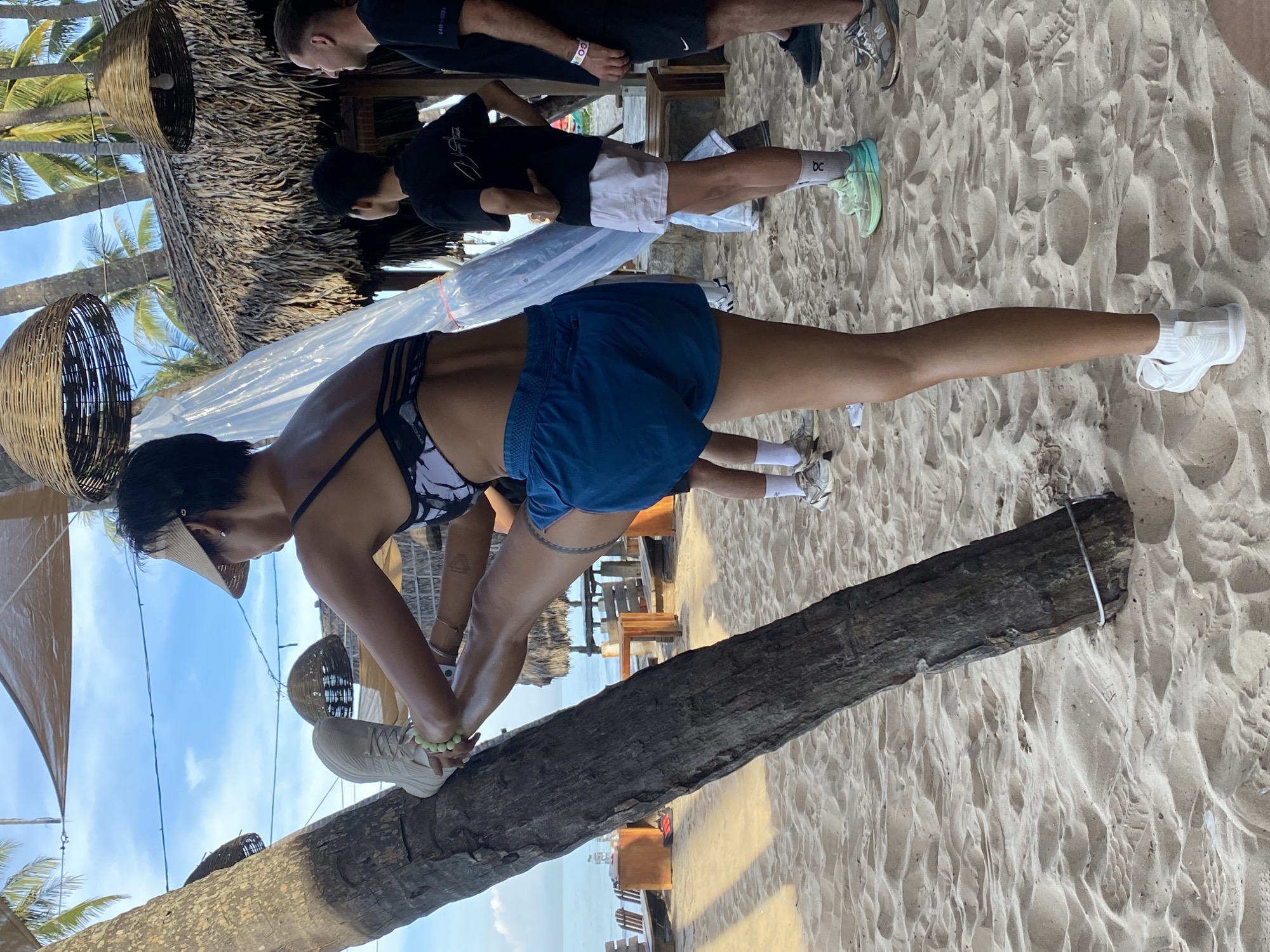 The image shows a woman in athletic wear stretching her leg against a palm tree on a sandy beach, likely in Mũi Né, Vietnam. She is wearing a black and white patterned sports bra, blue athletic shorts, white socks, and white athletic shoes. Her left leg is extended upwards and bent at the knee, supported by the tree trunk. She is also wearing a tan visor.
Several other individuals are visible in the background, also on the beach. To her right, a person wearing a black t-shirt and light-colored shorts is standing with their hand on their hip, looking away. Further to the right, another person in a black t-shirt and dark shorts is visible from the chest down. In the middle ground, behind the woman, another individual in black athletic wear and light shorts is visible.
The environment consists of a sandy beach with footprints and ripples from the tide. In the background, there are palm trees, thatched roofs of beachfront structures, and evidence of a relaxed, tropical atmosphere. The sky is blue with scattered clouds. The overall impression is that of an outdoor fitness activity taking place in a resort or recreational area along the coast.