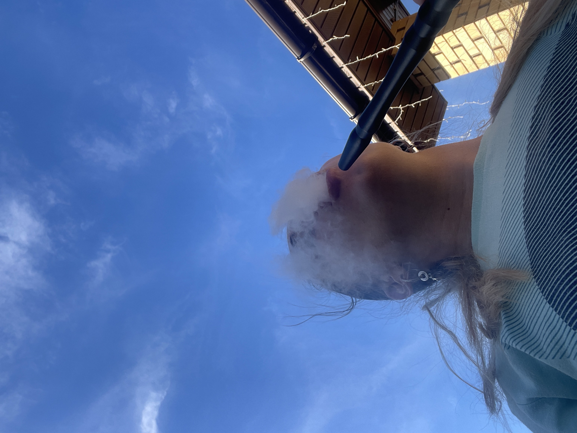 A low-angle shot captures a person exhaling smoke while holding a black hookah hose to their mouth. The person has fair skin and light brown hair, and they are wearing a striped shirt. Above the person, there is a dark-colored roof structure adorned with string lights. The sky is a clear blue with sparse clouds. The location is Chaikovsky, Russia.