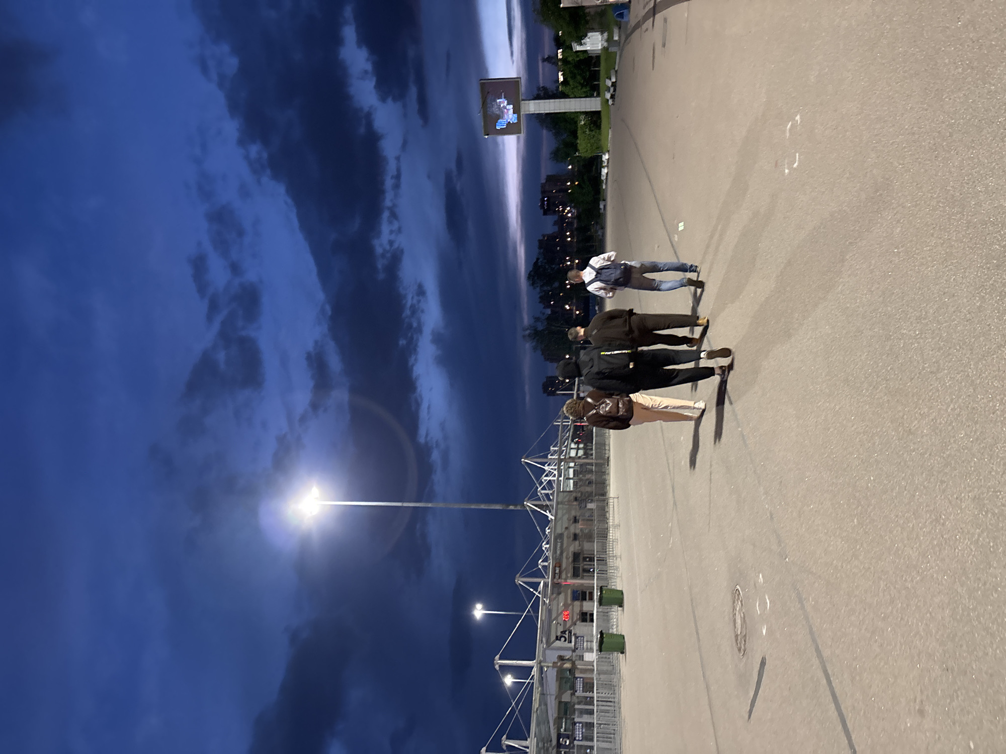 This image depicts an outdoor scene at twilight or early evening in Saint Petersburg, Russia. The sky is a dramatic dark blue, heavily laden with clouds, indicating either dusk or predawn.
The primary subjects are four individuals present on a wide, paved surface, likely a pedestrian walkway or plaza. One person, wearing blue jeans and a light-colored top with a backpack, walks away from the viewer. Behind them, two other individuals are dressed in dark clothing, and a fourth person is partially visible near them. They appear to be casually moving or standing in the area.
To the left, a bright streetlight illuminates the scene, casting a strong glow and a distinct lens flare against the dark sky. Further along the paved area, a complex of metal fences and modular structures, typical of stadium entrances or event barriers, stretches into the background. On one of these structures, a red digital display shows "5A" and some smaller numbers below, possibly "656," likely designating a gate or section number for a large venue.
In the distance, the silhouettes of city buildings are visible against the horizon. A large, rectangular billboard or screen is prominently displayed above the distant landscape, showing an illuminated graphic or video, though its specific content is not discernible. The overall setting suggests the perimeter or entrance area of a stadium or a large public event facility.