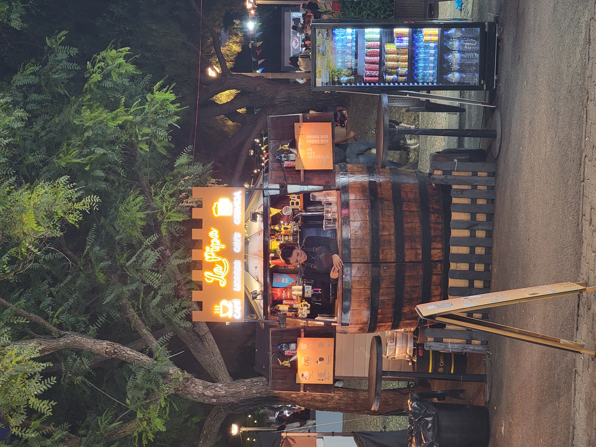 This is an outdoor night scene at a food and drink stall in Quarteira, Portugal. The stall is built into a large wooden barrel, with a canopy displaying the name "La Pipa" in neon lettering. Behind the counter, a woman wearing a dark apron and glasses is preparing drinks. The stall is stocked with various beverages, including sodas and bottled water. To the right of the stall is a refrigerated display case full of drinks. In the foreground, there are two small tables, a trash can, and an A-frame sign. The background features trees, people, and string lights, suggesting a lively atmosphere. The ground is paved with cobblestones.