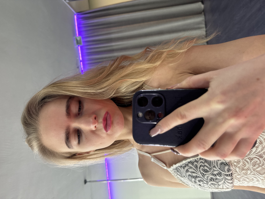 A young adult female is depicted in a mirror selfie, holding a dark-colored smartphone with her right hand. She has long blonde hair, styled in waves, and wears a light-colored, possibly off-white, lace top with thin shoulder straps. Her gaze is directed downwards towards the phone screen, and her lips are pursed, covered in dark pink lipstick. Her eyebrows are prominent. Her right hand, featuring manicured nails, obscures part of her torso and holds the phone, reflecting a partial image of her face in the phone's camera lenses. The setting, reflected behind her, is an indoor space within the city of Unknown, Unknown, likely a studio or fitness room. Visible elements include a metallic pole on the far left, a light-colored ceiling, and a purple LED strip light mounted above dark grey curtains in the background. The floor appears dark.