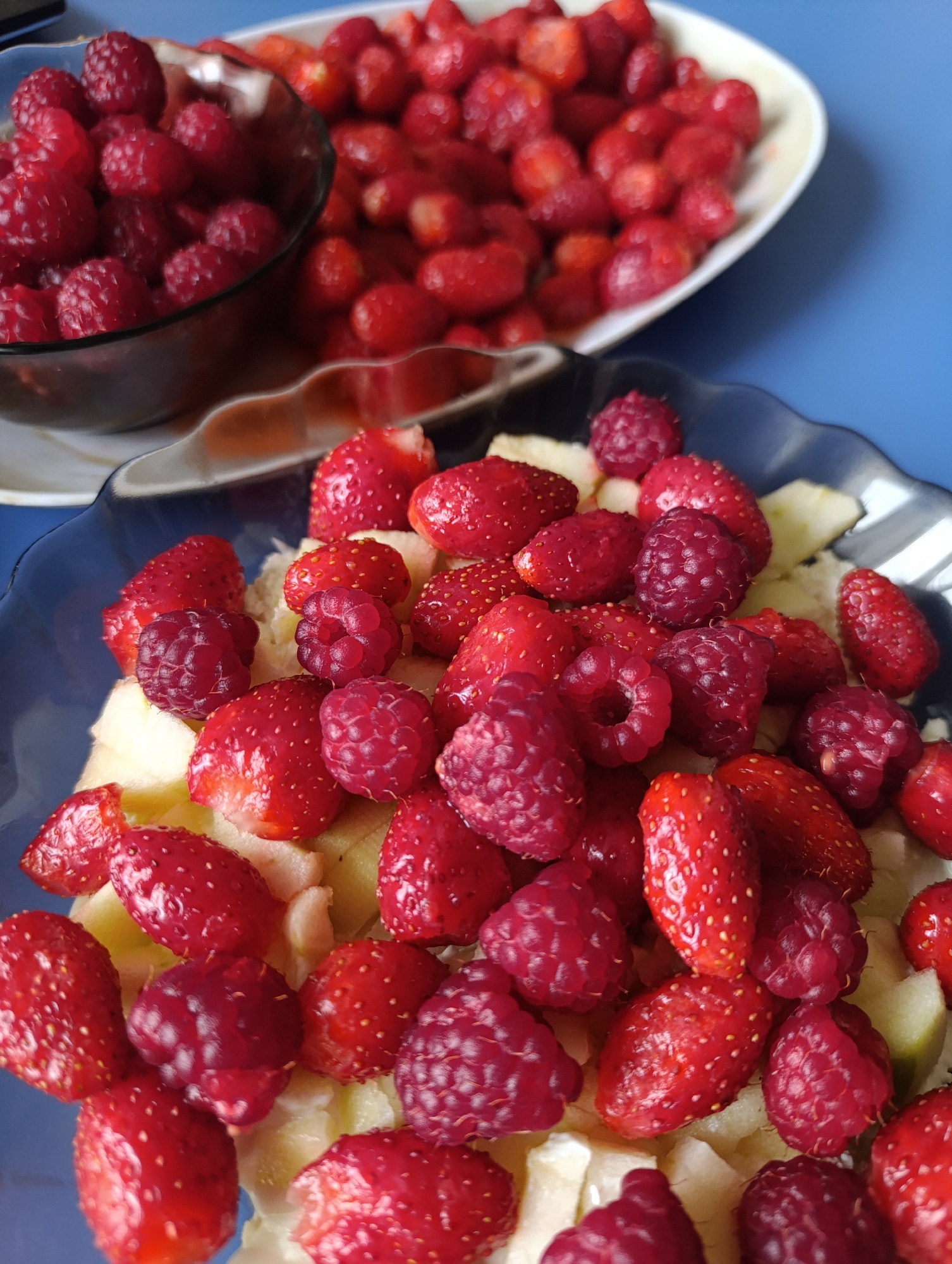 A close-up, high-angle shot shows a bowl filled with fresh raspberries and strawberries sitting on a blue surface. In the background, a white plate overflows with more of the same red berries. The raspberries in the bowl are plump and a deep red, while the strawberries are bright red with visible seeds. The white plate is filled with strawberries and raspberries, and the blue surface extends to the edges of the frame.