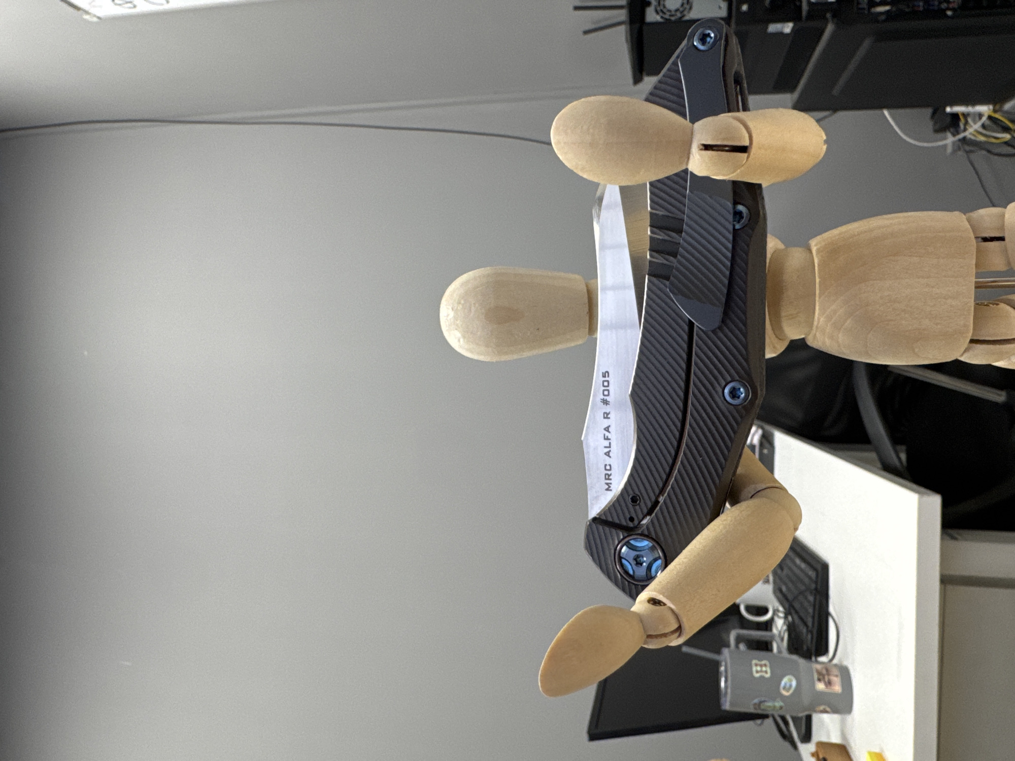 A wooden mannequin is holding a closed pocket knife. The knife has a dark carbon fiber handle and a silver blade with the inscription "MRC ALFA R #005". The mannequin appears to be positioned on a white desk or table, with a computer monitor and various electronic devices visible in the background. The background also includes a grey wall with a wire running along it. The location is not possible to identify from the provided image.