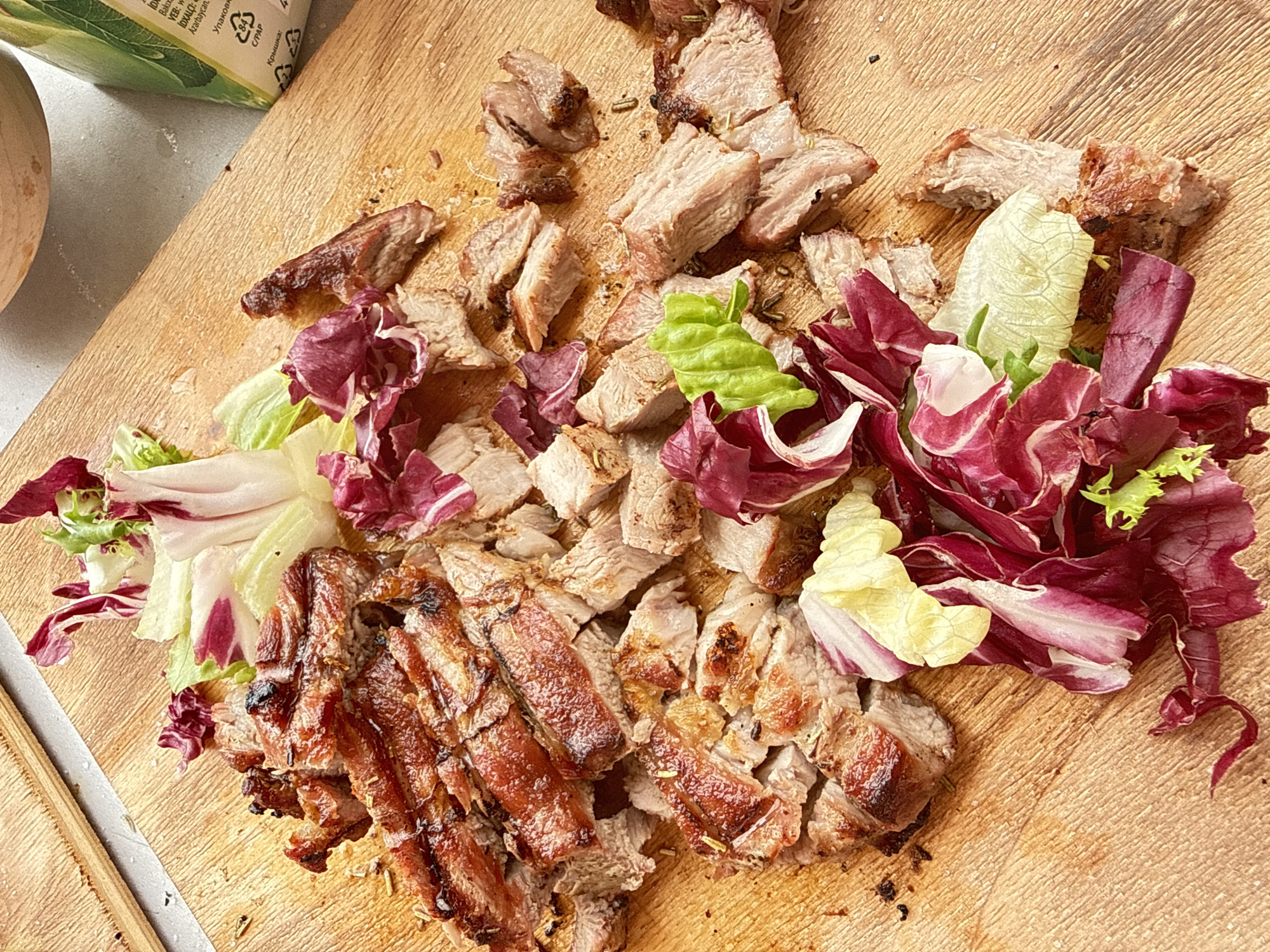 The image shows a wooden cutting board on a grey surface with grilled meat and lettuce on it. The meat appears to be cut into cubes and strips. There are different types of lettuce, including a red variety. The scene appears to be food preparation. There is a container in the upper left corner. It is not possible to determine a location, such as the city of Chaikovsky, Russia, from the information in the image.