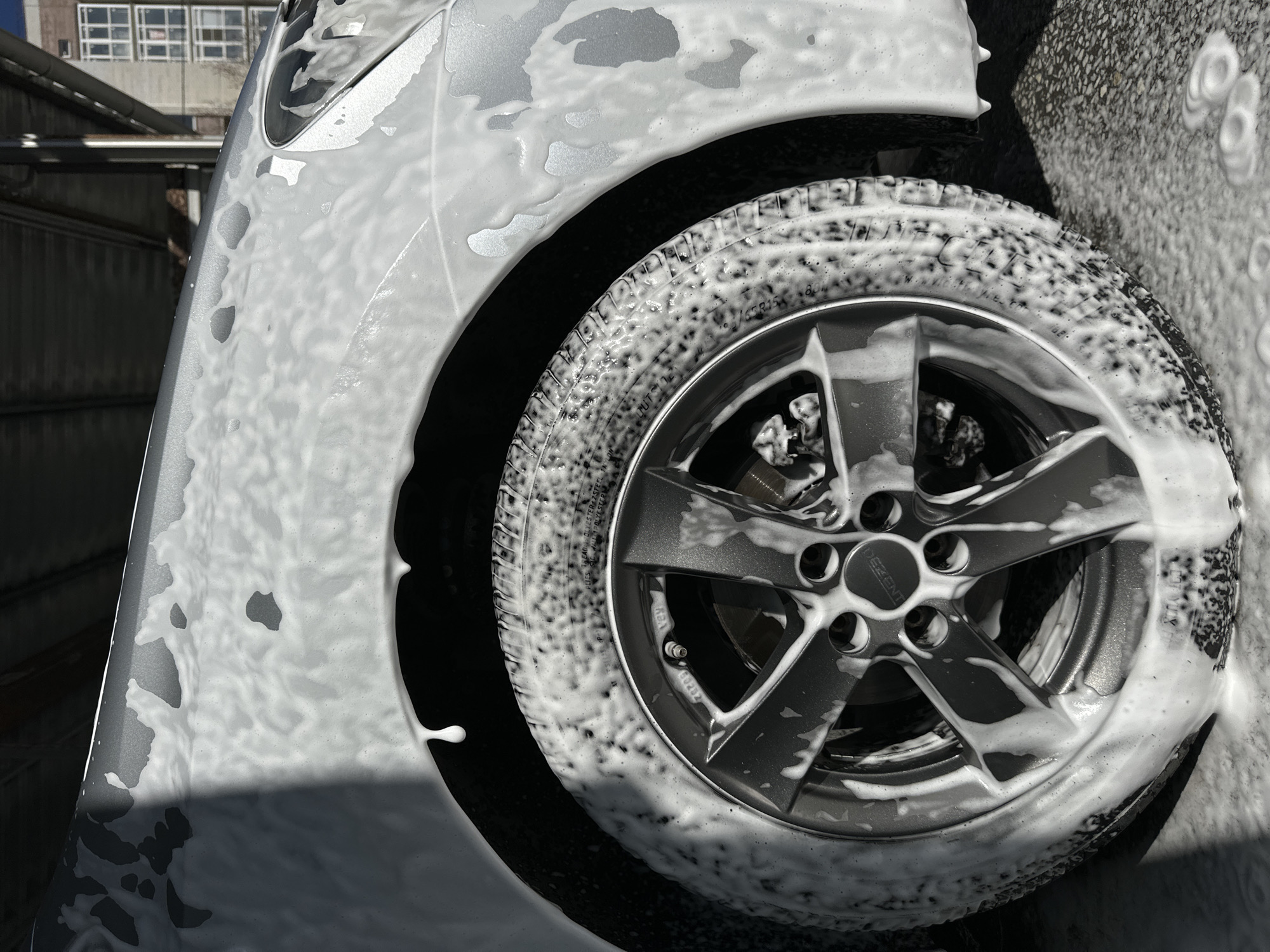 A close-up shot depicts the front portion of a silver-colored car being washed. The vehicle's front fender and a tire are heavily coated in white soap or cleaning foam. The tire has a five-spoke alloy wheel. The make "DEZENT" is visible on the wheel's center cap. The photograph highlights the cleaning process, with the foam obscuring some details of the car. The image does not depict any people or specific objects related to Novoaleksandrovka, Russia.