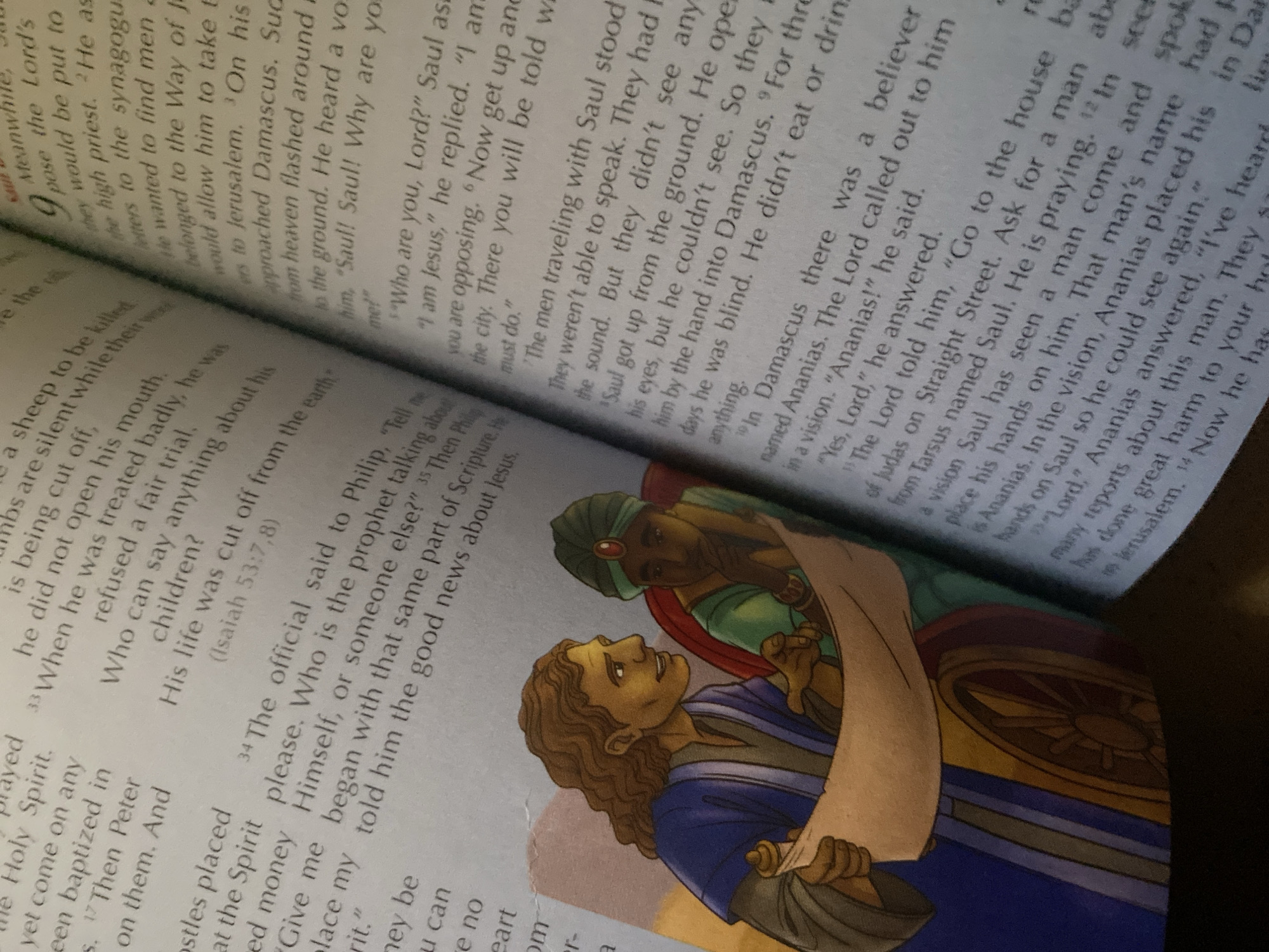 The image shows an open book with text and an illustration. The illustration depicts two men looking at a scroll. One man on the left has curly brown hair and is wearing a blue and purple robe. He is pointing at the scroll with his left hand. The man on the right has dark skin and is wearing a green robe and a turban. He is looking at the scroll with his chin resting on his hand. The scroll itself appears to be a map or document. The text in the book appears to be from the Bible. The scene is set against a blurred background. There is no visible indication of the city of Jauro Yinu, Nigeria.