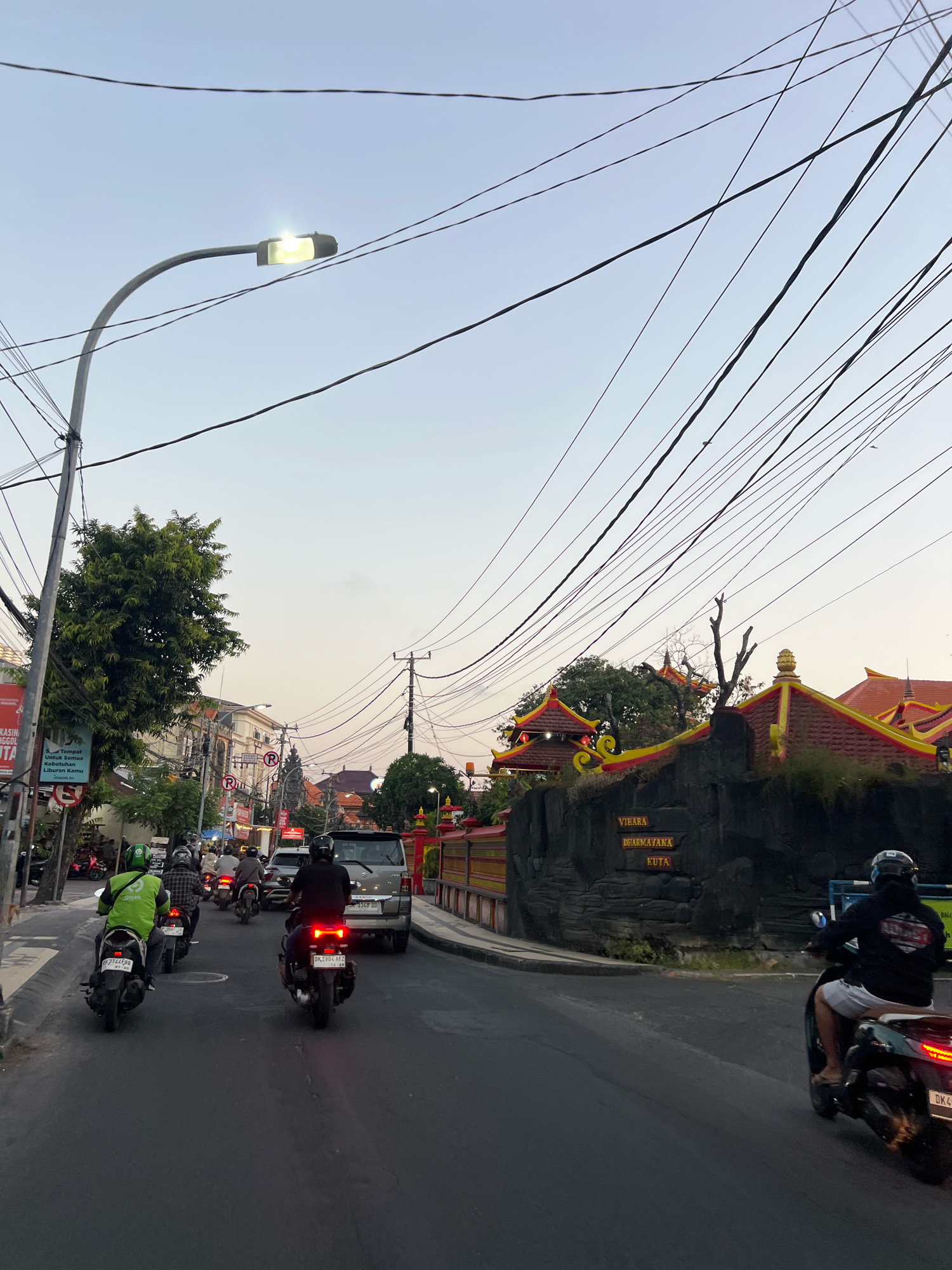 This image captures a street scene in Kuta, Indonesia, likely during the late afternoon or early evening. The sky is a pale, hazy orange, indicating either sunrise or sunset. A street lamp is illuminated on the left side of the image, casting a bright light. The street is paved and appears to be a busy thoroughfare, with several motorcycles and a van on the road.
In the foreground, a person on a motorcycle wearing a dark jacket and shorts is moving away from the viewer. To their left, a motorcycle with a rider wearing a green jacket, possibly a delivery driver, is also moving forward. Further back, other motorcycles and a silver van are visible in traffic.
The right side of the road features a distinctive building with a dark, rock-like facade adorned with colorful, traditional Indonesian architectural elements, including red and yellow tiled roofs. A sign on the facade reads "VIHARA DHARMAYANA KUTA," indicating it is likely a Buddhist temple or monastery. Lush green trees are present on the left side of the street, bordering buildings that appear to be shops or residences. Overhead, a complex network of electrical wires crisscrosses the sky, a common sight in many urban environments. The overall atmosphere suggests a typical day in a bustling Indonesian city.