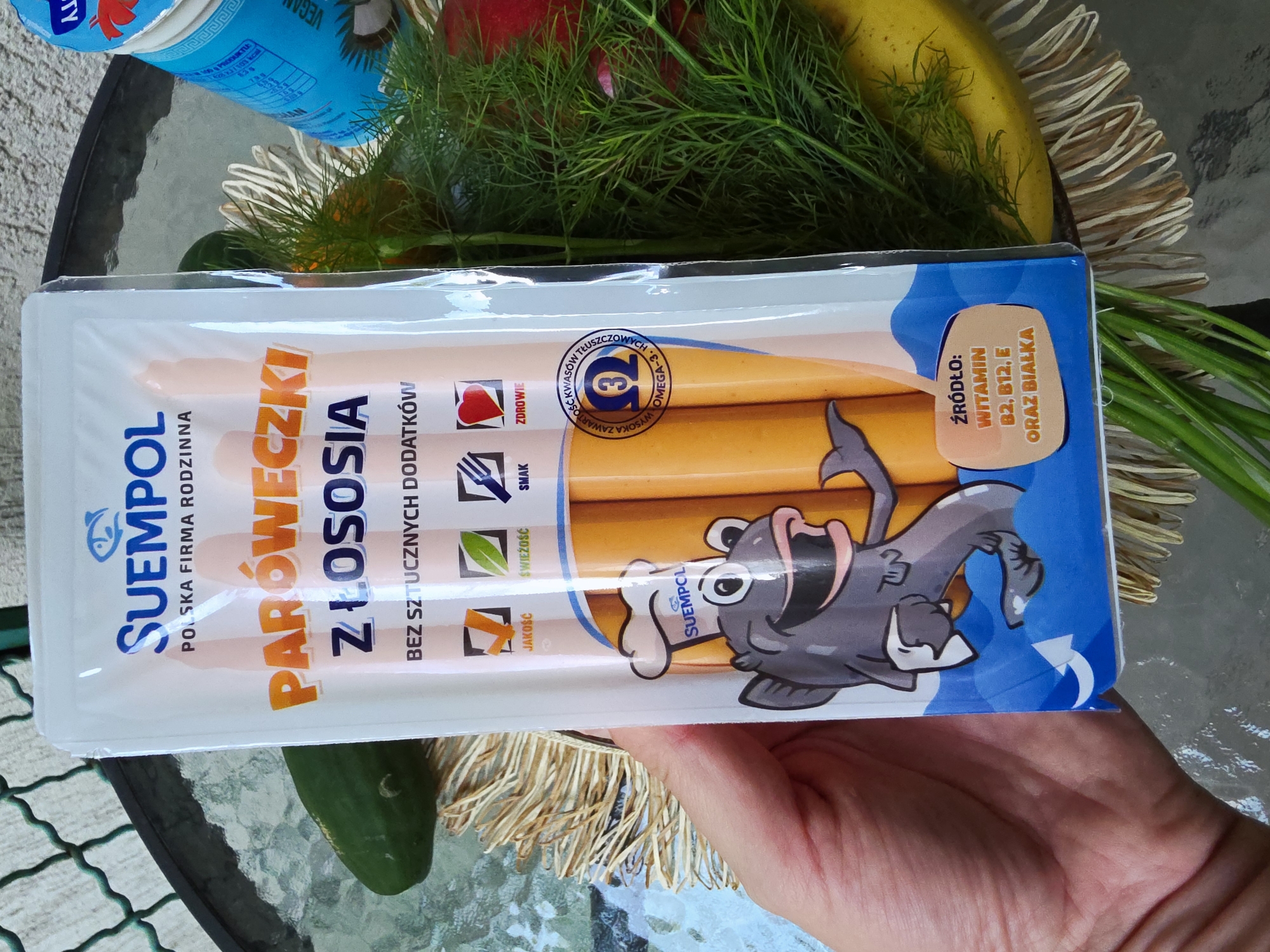 A person's hand holds a package of "SUEMPOL PARÓWECZKI Z ŁOSOSIA" (Salmon Sausages). The white and blue packaging features a cartoon fish wearing a chef's hat. The brand "SUEMPOL" is described as a "POLSKA FIRMA RODZINNA" (Polish Family Company).
Prominently displayed text on the package highlights "BEZ SZTUCZNYCH DODATKÓW" (No Artificial Additives). Several icons indicate product attributes: "JAKOŚĆ" (Quality), "ŚWIEŻOŚĆ" (Freshness), "SMAK" (Taste), and "ZDROWIE" (Health). A circular emblem states "WYSOKA ZAWARTOŚĆ KWASÓW TŁUSZCZOWYCH OMEGA-3" (High content of Omega-3 fatty acids). Another section indicates the product is a "ŹRÓDŁO: WITAMIN B2, B12, E ORAZ BIAŁKA" (Source of: Vitamins B2, B12, E and Protein).
The package contains several light orange, cylindrical sausages. In the background, on what appears to be a glass outdoor table, are various groceries. These include fresh dill, a yellow banana, red vegetables (possibly radishes), and a blue container, partially visible, which has text like "VEGAN" and nutritional information. The setting suggests a casual, possibly outdoor, environment with fresh produce. The image is well-lit, indicating daytime.