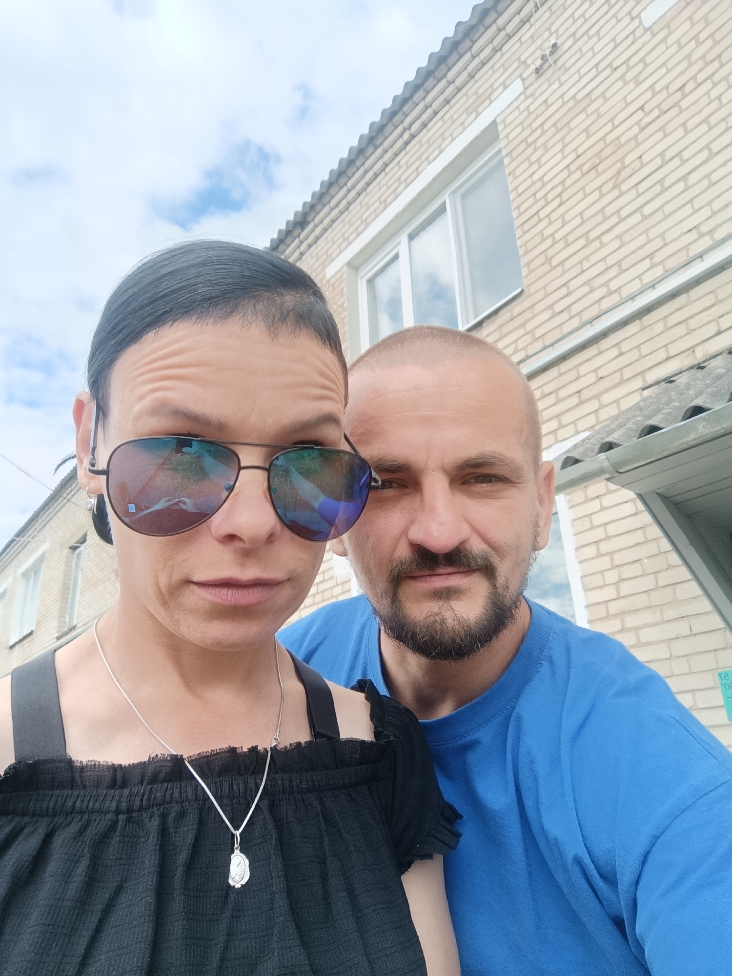 This is a selfie of a man and a woman in front of a brick building. The woman is on the left, wearing sunglasses, a black off-the-shoulder top, and a silver necklace. She has dark hair pulled back in a ponytail. The man is on the right, wearing a blue t-shirt and has short hair and a beard. The building behind them is made of light-colored bricks and has a gray roof. There are windows visible on the building. The sky is cloudy. The image appears to be taken during the daytime, likely outdoors. The subjects' facial expressions are neutral. There is no visible text in the image.