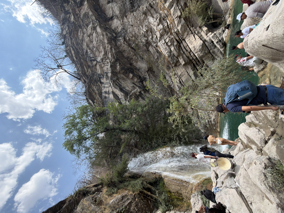 This image depicts a natural recreational area in San Miguel Atlapulco, Mexico, centered around a waterfall and a turquoise pool. The scene is dominated by steep, rocky cliffs on both sides, with the right cliff face exhibiting prominent horizontal stratification. The sky above is clear blue with scattered white cumulus clouds.
On the left, a waterfall cascades down a verdant, rocky cliff into a tranquil, teal-colored pool below. The surrounding vegetation includes both lush green trees and shrubs near the waterfall, and sparser, bare-branched trees on the higher parts of the cliffs, particularly visible on the right slope.
Approximately ten to twelve individuals are present within the scene. In the foreground, positioned to the right, a man wearing a dark blue t-shirt, blue jeans, a black baseball cap, and a blue backpack faces away from the camera, looking towards the water. On the rocky bank near the base of the waterfall, a person with long dark hair, dressed in a white t-shirt and dark pants, stands next to a large, light-colored, circular object resembling a reflector or a drum. Adjacent to them, a woman in a white two-piece swimsuit stands on a rock, facing the water with her back to the camera. Further to the right, several individuals are partially submerged in the turquoise water or seated on the surrounding rocks, engaged in leisure activities. Their attire includes t-shirts, caps, and swimwear. The immediate ground consists of large, light-colored boulders and smaller rocks.
