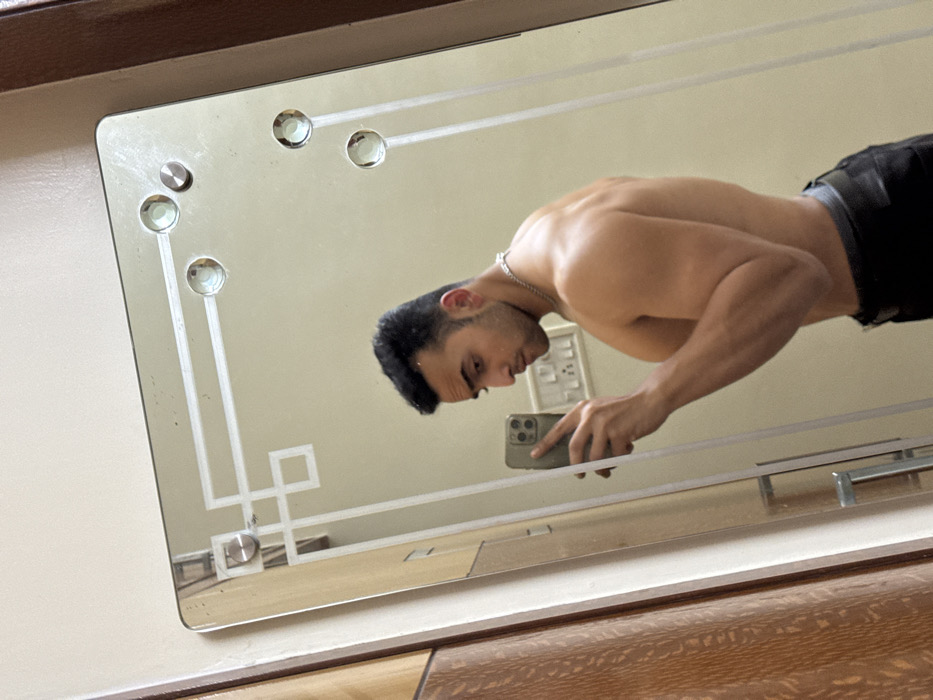 A shirtless man takes a selfie in a rectangular mirror with a geometric design and circular accents. He holds a smartphone in his hand, his face partially visible in the reflection. He wears black pants. The mirror hangs against a light-colored wall, and part of a wooden surface is visible at the bottom of the image.