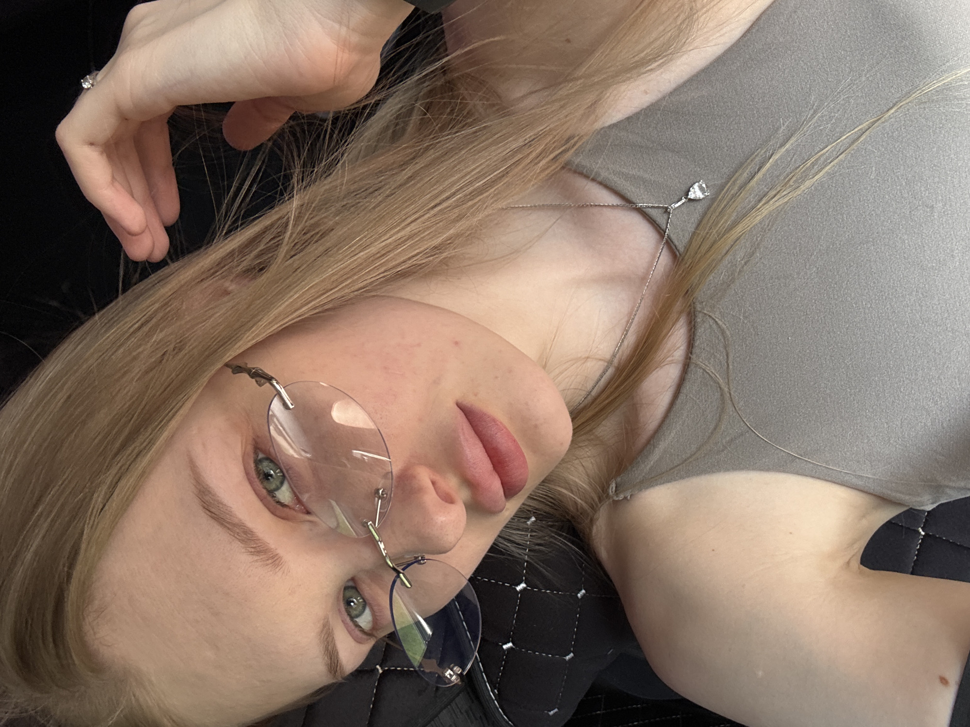 A woman with light blonde hair, wearing rimless glasses and a gray top, lies on a black quilted surface. She is facing the camera with her head tilted. A silver necklace with a small pendant is visible. A hand with a ring is partially visible in the upper left. The location in the city of Perm, Russia is not evident in the image.
