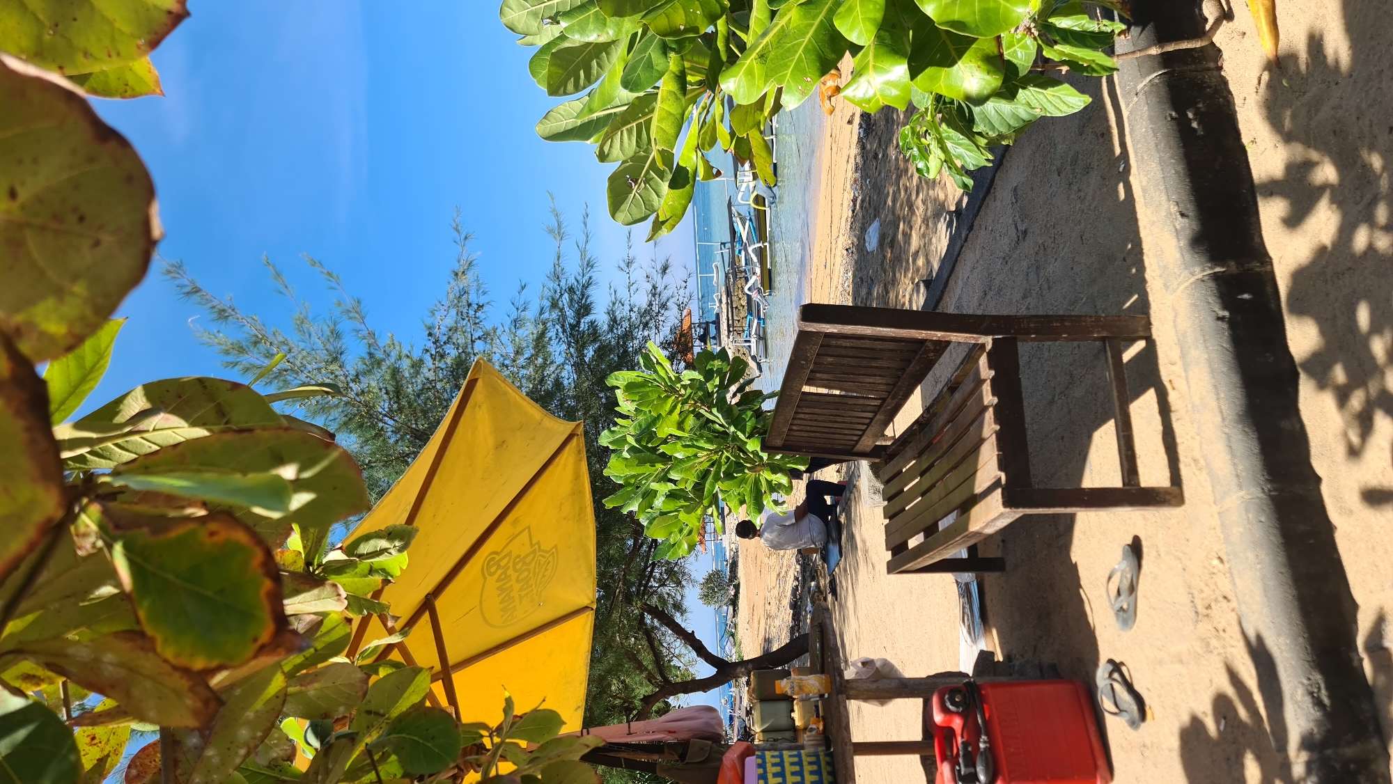 A man sits on the sand at a beach in Denpasar, Indonesia, facing away from the camera. He is wearing a white t-shirt and dark pants. In front of him is a wooden bench, and to his left, a small table with various items, including a red fuel canister, bottles, and a yellow umbrella providing shade. The beach extends to the water, where several boats are moored. Lush green foliage frames the scene from the foreground and mid-ground, with a clear blue sky overhead. The overall impression is one of a tranquil beachside setting.