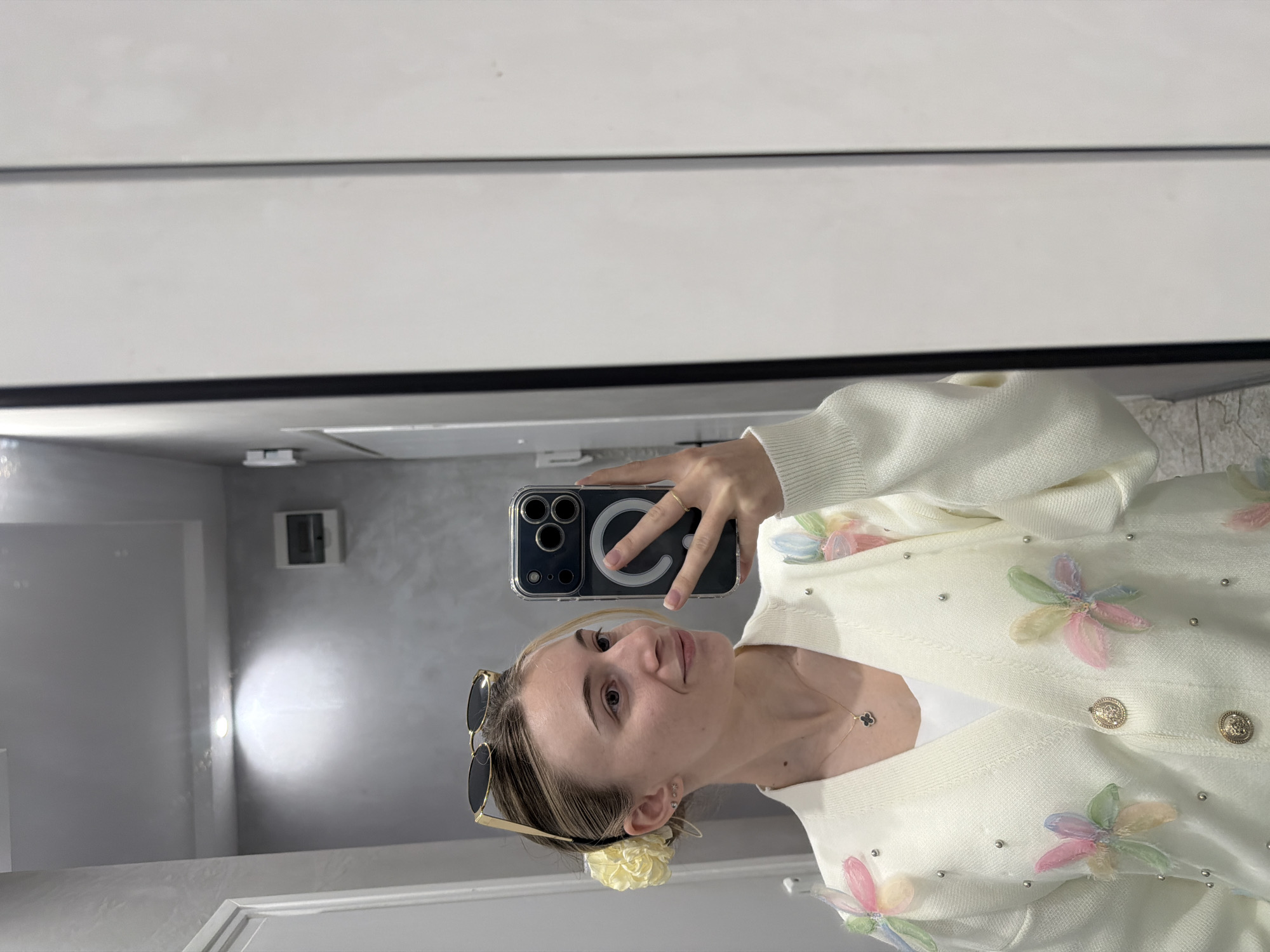 The image appears to be a mirror selfie taken indoors, with a woman in the foreground. She is holding a smartphone up to the mirror, capturing her reflection. She is wearing a white cardigan decorated with floral appliques and small pearl-like embellishments. She has sunglasses perched on her head and a light-colored flower in her hair. A necklace is visible around her neck.
The background is a blur of an interior space, possibly a bathroom or dressing room. There are white cabinets and walls, along with a mirror.
There is insufficient information in the image to determine if it was taken in Plovdiv, Bulgaria. No specific landmarks, architecture, or other environmental features are visible that would indicate a particular location.