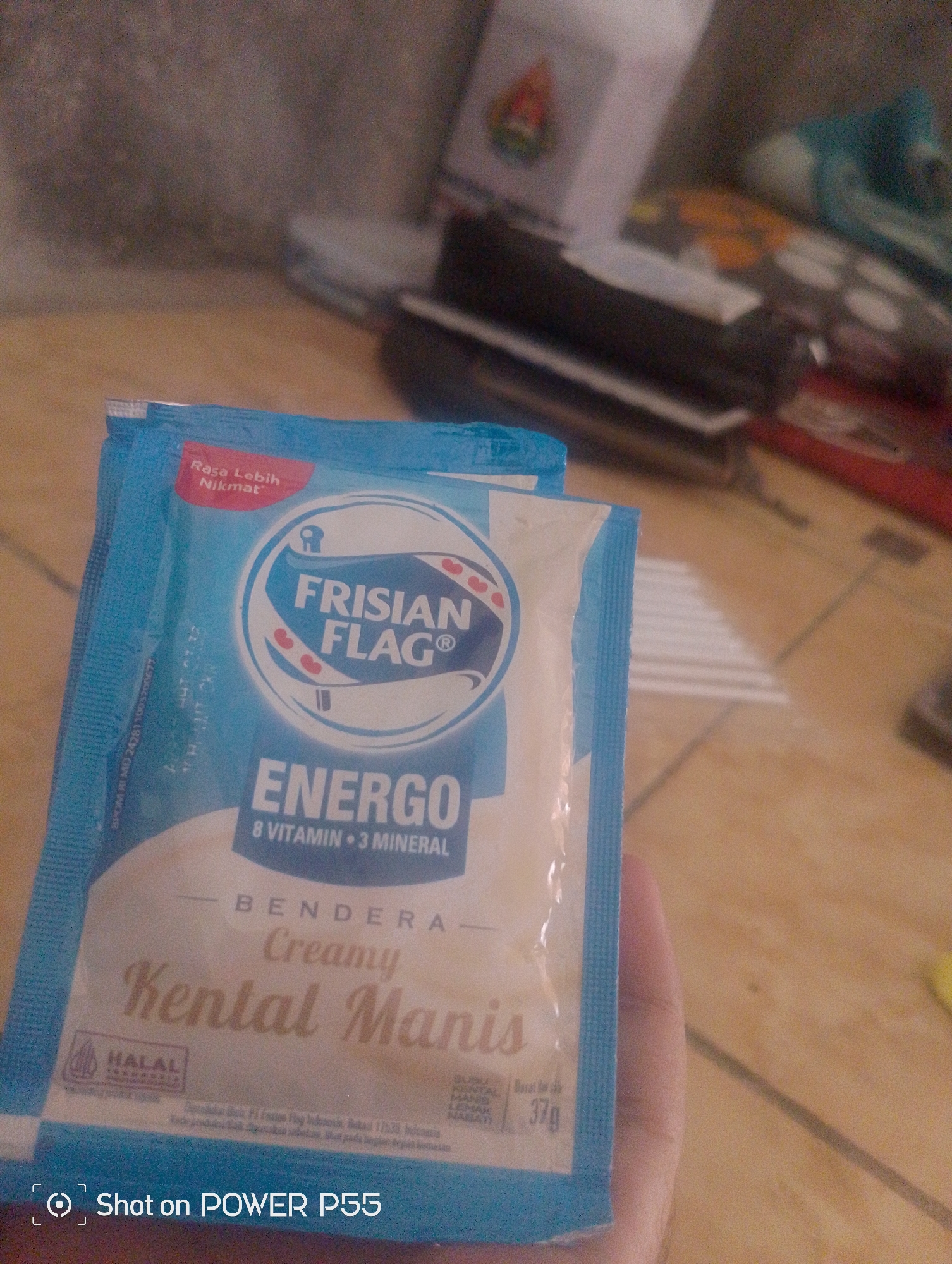 The image shows a close-up of a Frisian Flag Energ O "Bendera Creamy Kental Manis" sachet. The sachet is blue and white with red accents. The Frisian Flag logo is visible, as is text indicating "8 VITAMIN • 3 MINERAL". A HALAL certification is also present. The sachet is being held by a hand, partially visible at the bottom right of the frame. The background is blurred and appears to be a wooden surface with some out-of-focus objects. The image is a photograph taken with a "POWER P55" device. No specific location in Lebaksiu, Indonesia is identifiable from the image.
