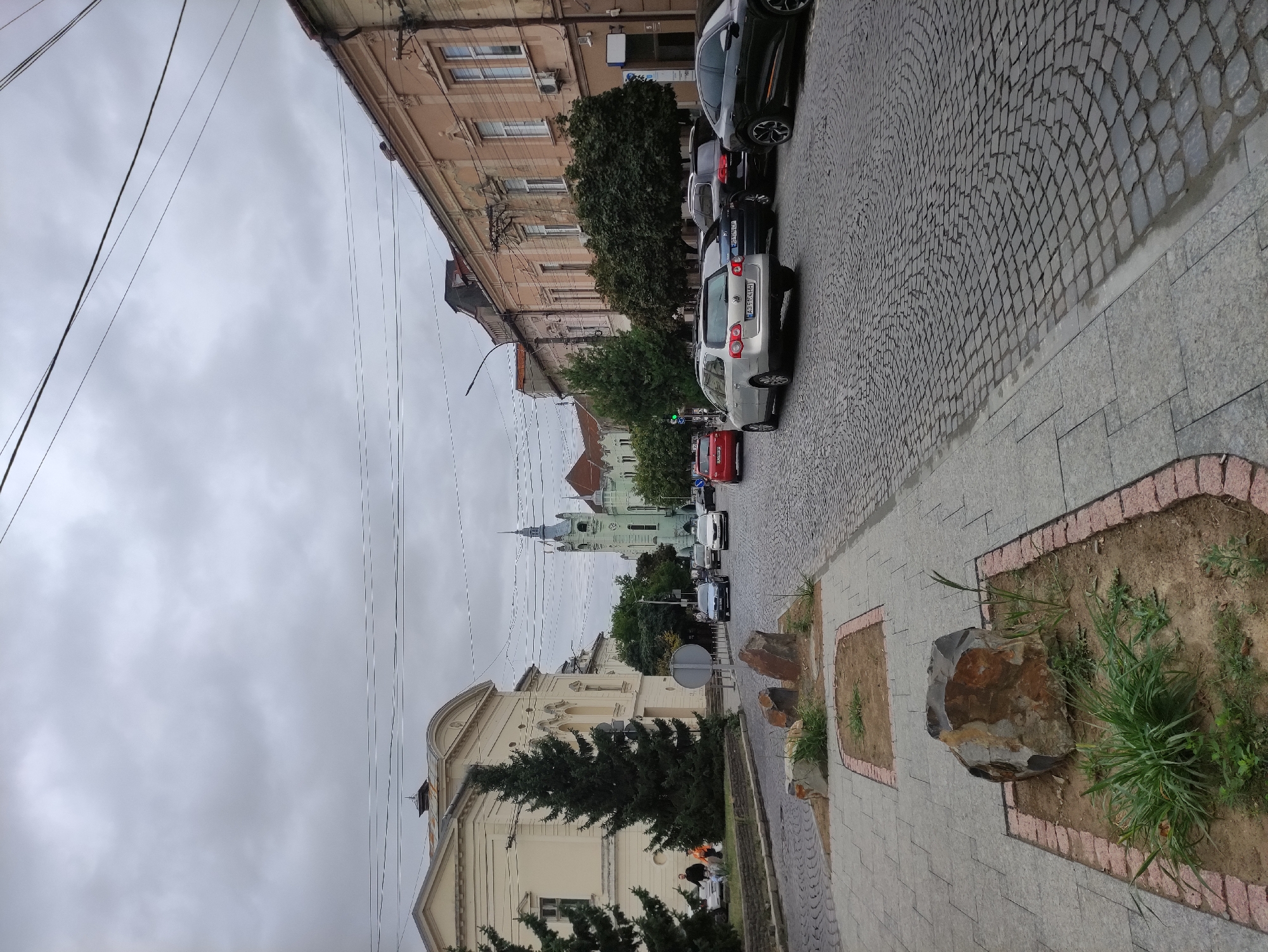 The image shows a street scene in Mukachevo, Ukraine, likely near the Drama Theater and the Greek Catholic Cathedral, judging by the architectural styles and prominent tower visible. The sky is overcast. The street is paved with cobblestones, and a sidewalk made of large gray rectangular tiles runs along the left side of the road. There are several cars on the street, including a silver station wagon in the foreground, a black SUV behind it, and a black coupe further down the road. Other vehicles are visible further up the street, including a red car. On the left side of the street, a light-colored building with arched windows is visible, with two evergreen trees in front of it. People are walking on the sidewalk in front of this building. A distinctive green tower with a clock is prominent in the background, a landmark in Mukachevo. In the foreground, on the tiled sidewalk, there are several planters with dirt, rocks, and sparse vegetation. The overall atmosphere is that of a typical European city street on a cloudy day.