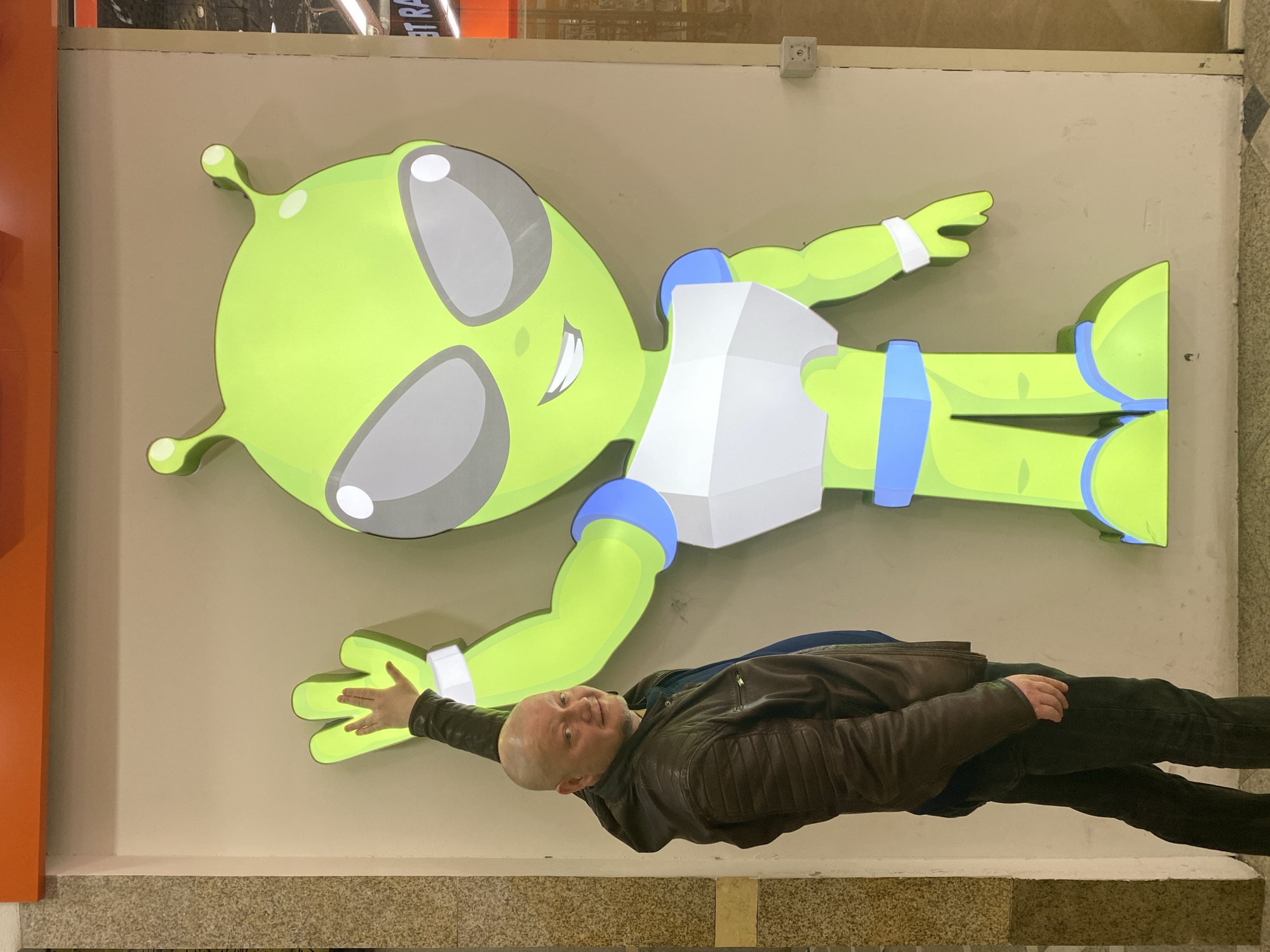 A man in a brown leather jacket and dark pants is lying on the floor beneath a large, cartoon-style alien figure displayed on a wall. The alien is green with large, gray-tinted eyes, blue accents on its wrists, waist, and ankles, and a white triangular garment. The man's right hand is extended toward the alien's hand. The wall is beige, and a strip of orange is visible on the left side of the frame. A light fixture and ventilation duct are visible near the ceiling. The floor is tiled. This scene is located in Yekaterinburg, Russia.