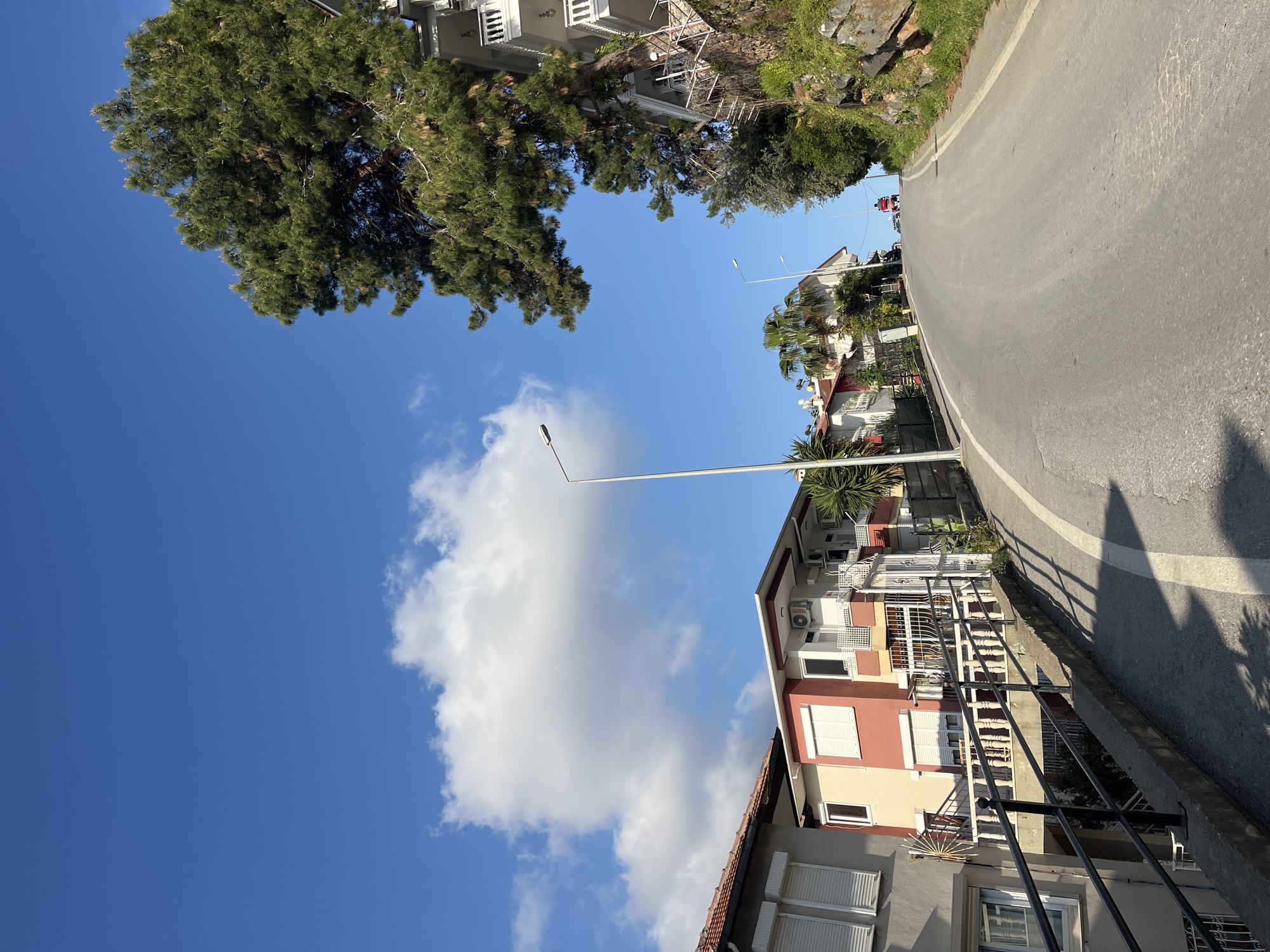 The image captures a sunny, clear day on a residential street in Alanya, Turkey. The scene is dominated by a vibrant blue sky with a few scattered white cumulus clouds.
On the left side of the frame, a large, lush green tree, likely a pine, extends its canopy prominently, partially framing the sky. Below and to the right of this tree, glimpses of other buildings and greenery suggest a built-up area.
The right side of the image features an asphalt road that gently curves upwards and away from the viewer. A solid white line marks the edge of the road, and dark, elongated shadows are cast across its surface and an adjacent concrete barrier. These shadows indicate the sun is high in the sky, likely mid-morning or early afternoon.
Alongside the road on the right are multi-story residential buildings. One prominent building has a light yellow lower facade with a reddish-brown upper section, featuring balconies with dark wrought-iron railings and white window shutters. Air conditioning units are mounted on its exterior, with the brand "VESTEL" clearly visible on one. Another building with a grey facade and white shutters is partially visible in the foreground. Palm trees and other Mediterranean foliage are scattered around the properties. A tall, slender streetlight pole stands in the midground, its lamp head extending towards the center of the sky.
In the distance, a small red vehicle is partially visible on the road, suggesting ongoing, albeit light, traffic. The overall scene depicts a typical warm, Mediterranean urban landscape.