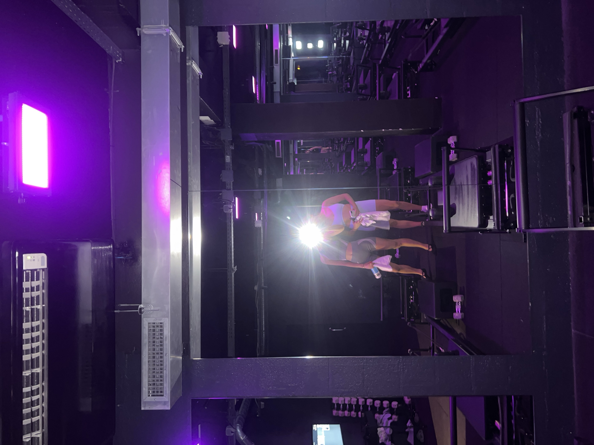 The image shows a group fitness class taking place in a gym. The room is dimly lit with purple lighting, creating a vibrant and energetic atmosphere. A bright light, possibly from a projector or a large lamp, illuminates the center of the room, casting a glare. Two individuals are visible in the reflection of a mirror. One is wearing a white tank top and grey shorts, holding a towel and a bottle of water. The other is wearing a light-colored athletic outfit, also holding a towel. They appear to be engaged in a workout, possibly during a break or instruction.
The gym equipment is visible in the background, including weight machines and racks of dumbbells. The overall setting suggests a modern fitness studio. The scene is indoors, and there are no indicators of time of day or weather. The purple lighting and reflections create a sense of a high-energy, possibly late-night or evening, workout session. The location context of Paris, France, does not offer specific visual cues in this image. No visible text is present.