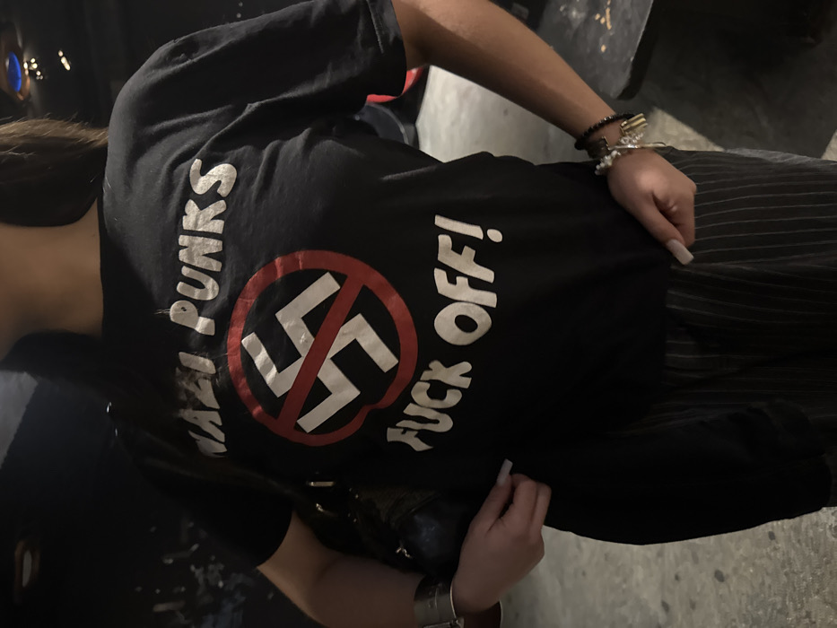 The scene is situated in Paris, France. A person, identified from the mid-torso up, stands indoors. The subject wears a black short-sleeved t-shirt with white text reading "NAZI PUNKS" above a red circle containing a diagonal line intersecting a white swastika, and "FUCK OFF!" below. The person's hair is dark and falls over their shoulders. Dark, vertically striped pants are visible from the waist down. On the left wrist, a wide, silver-toned cuff bracelet is present, and a dark bag with a strap is slung over the left shoulder. The right wrist displays multiple beaded bracelets. The subject's hands, featuring long, light-colored fingernails, are positioned near the waistline, with the left hand touching the t-shirt's hem and the right hand resting at the hip. The background is dark and out of focus, suggesting a dimly lit indoor environment, possibly a venue, with a dark floor.