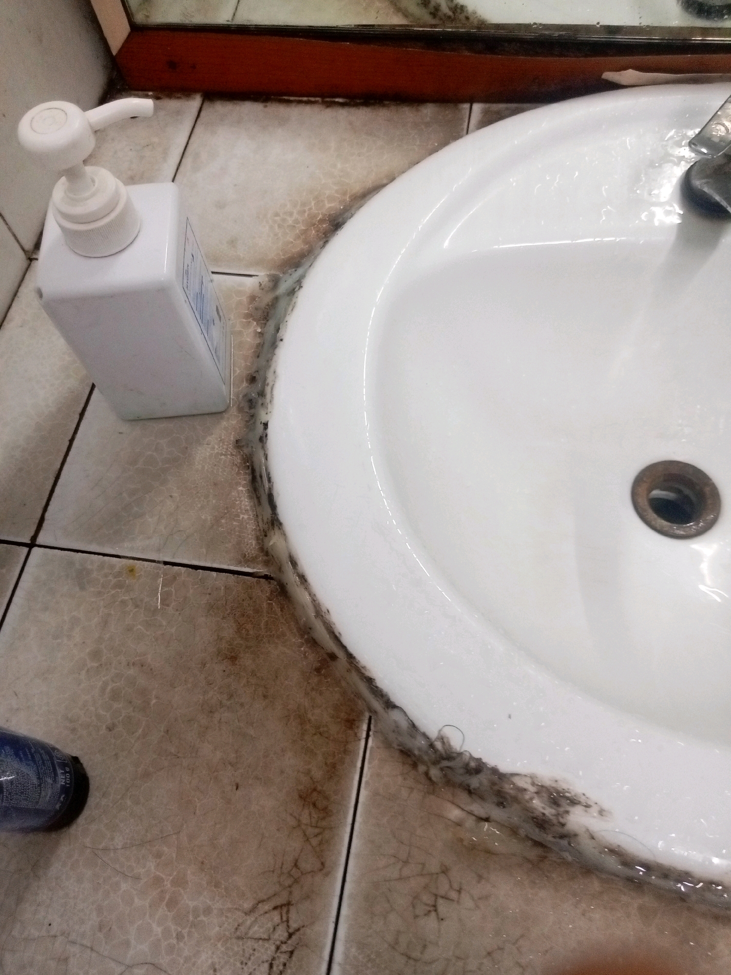 The image depicts a close-up, high-angle view of a dirty bathroom sink area. A white pump bottle dispenser is situated on the tiled floor to the left of the sink. The tiles themselves are light-colored with a cracked pattern and appear stained and grimy, particularly along the grout lines and around the base of the sink. Significant black mold or mildew is visible along the edge of the white ceramic sink, mixed with what appears to be hair and debris. The sink basin itself is white and reflects light, with a metallic drain visible in the lower right portion of the sink. Water droplets are present in the sink. A dark-colored object, possibly another bottle, is partially visible in the bottom left corner of the frame. The overall impression is one of neglect and poor hygiene. The location is stated to be Batam, Indonesia.