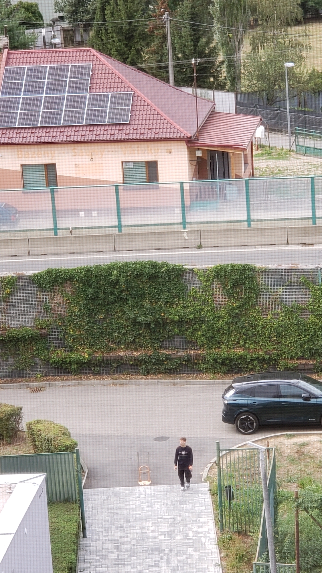 The image depicts an outdoor scene with a residential building featuring solar panels on its roof. A dark green SUV is parked on a paved area in front of a wall covered in ivy. A young man wearing a dark long-sleeved shirt and dark pants is walking on a paved walkway. The walkway leads to a set of stairs and appears to be an entrance to a building. The background includes trees and a fence. The location is identified as Prešov, Slovakia.