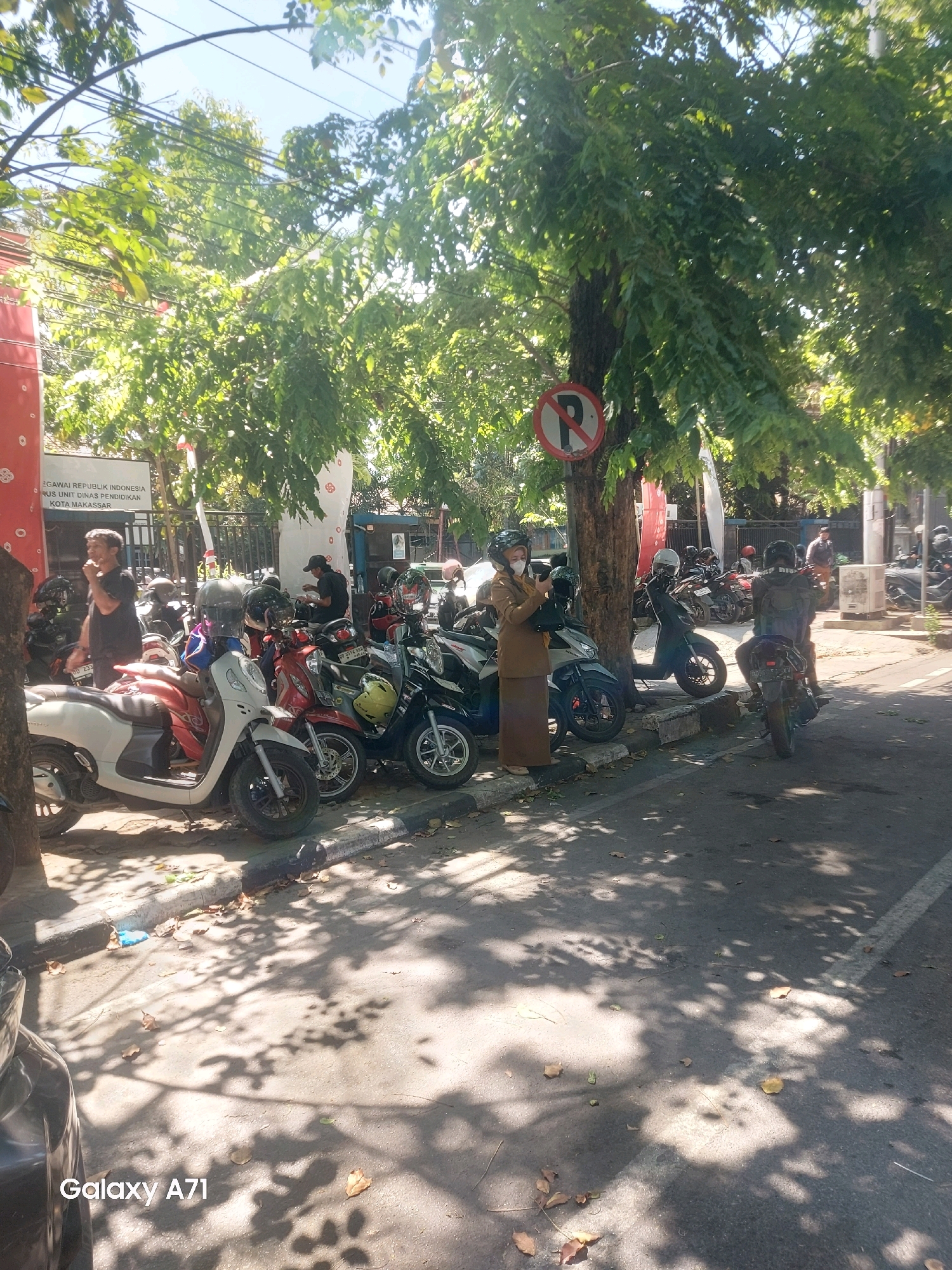 The image shows a sunny outdoor scene in Makassar, Indonesia, with a row of parked motorcycles lining a sidewalk in front of a building. A "no parking" sign is visible on a tree. Several people are present, some standing and others on or near motorcycles, suggesting a public area or parking lot. The environment is characterized by lush green trees casting shadows on the ground, indicating a warm, possibly humid, day. The building in the background appears to be a public institution with Indonesian text on its facade.