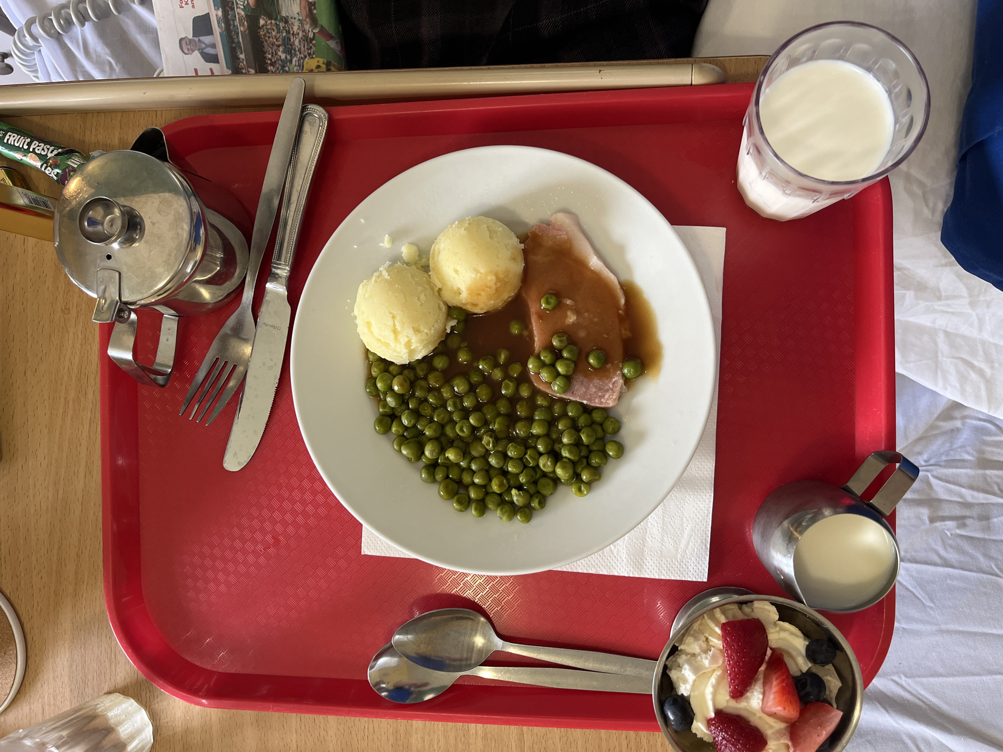 This is a top-down, eye-level, close-up, medium shot of a hospital meal tray placed on a red plastic tray. The meal consists of a white plate with mashed potatoes, peas in gravy, and a slice of ham. To the left of the plate, there are two spoons. To the left and bottom of the tray, there is a small metal bowl filled with whipped cream and berries, and a small metal jug containing milk. To the right and bottom of the tray, there is a clear glass filled with milk. In the background, on the right side of the tray, there is a glimpse of a newspaper and a silver teapot. The meal appears to be on a wooden surface, possibly a bedside table in a hospital room. The location is not Tyrrellspass, Ireland, but rather an unspecified hospital.