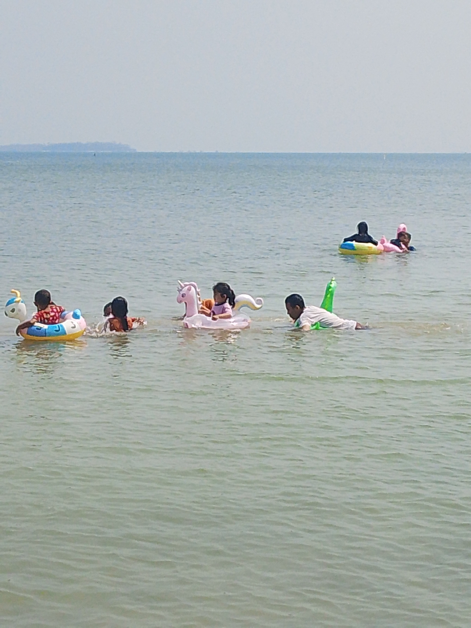 In the shallow, calm waters of the ocean near Pejagan, Indonesia, a group of children enjoys a sunny day. A young boy is positioned on the left side of the frame, sitting in a yellow and blue inflatable ring that has a duck design on the front. To his right, another child, mostly submerged, is also in the water, possibly playing. Further to the right, a girl sits atop a pink inflatable unicorn. In the middle ground, two children are in the water, with one appearing to be sitting on a yellow inflatable ring. On the far right, a child in a white shirt is swimming in the water, accompanied by a green inflatable toy. The horizon line is visible in the distance, with a hazy landmass appearing as a silhouette against the pale sky. The water is a muted green-blue, with gentle ripples on the surface.