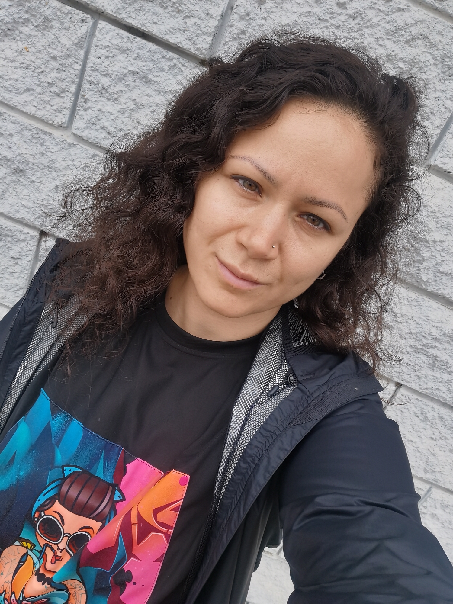 A mid-shot, slightly angled selfie shows a woman with curly dark brown hair, brown eyes, and a nose ring, looking at the camera. She is wearing a black t-shirt with a colorful graphic print on it and a black jacket with a gray mesh lining visible at the collar and zipper. The background is a light gray textured wall, possibly made of concrete blocks. The lighting suggests an outdoor setting on an overcast day. The woman's expression is neutral, with a slight smile. There are no other people or significant objects in the frame. The image does not contain any discernible elements that would indicate its location in Chaikovsky, Russia.