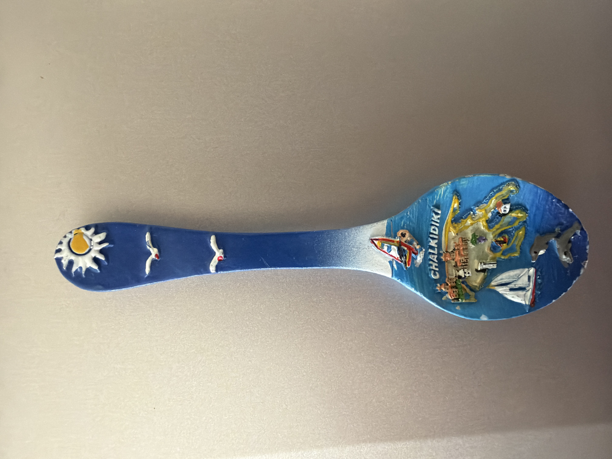 The image shows a spoon-shaped magnet, likely a souvenir. The top of the spoon features a stylized sun with rays and three white birds in flight against a blue background. The lower portion of the spoon depicts a scene of a coastal area with islands, buildings, a sailboat, a windsurfer, and two dolphins swimming. The text "CHALKIDIKI" is visible on the spoon. The background of the image is a plain, light-colored surface, possibly a refrigerator door. There is no indication of Pazardzhik, Bulgaria in the image.