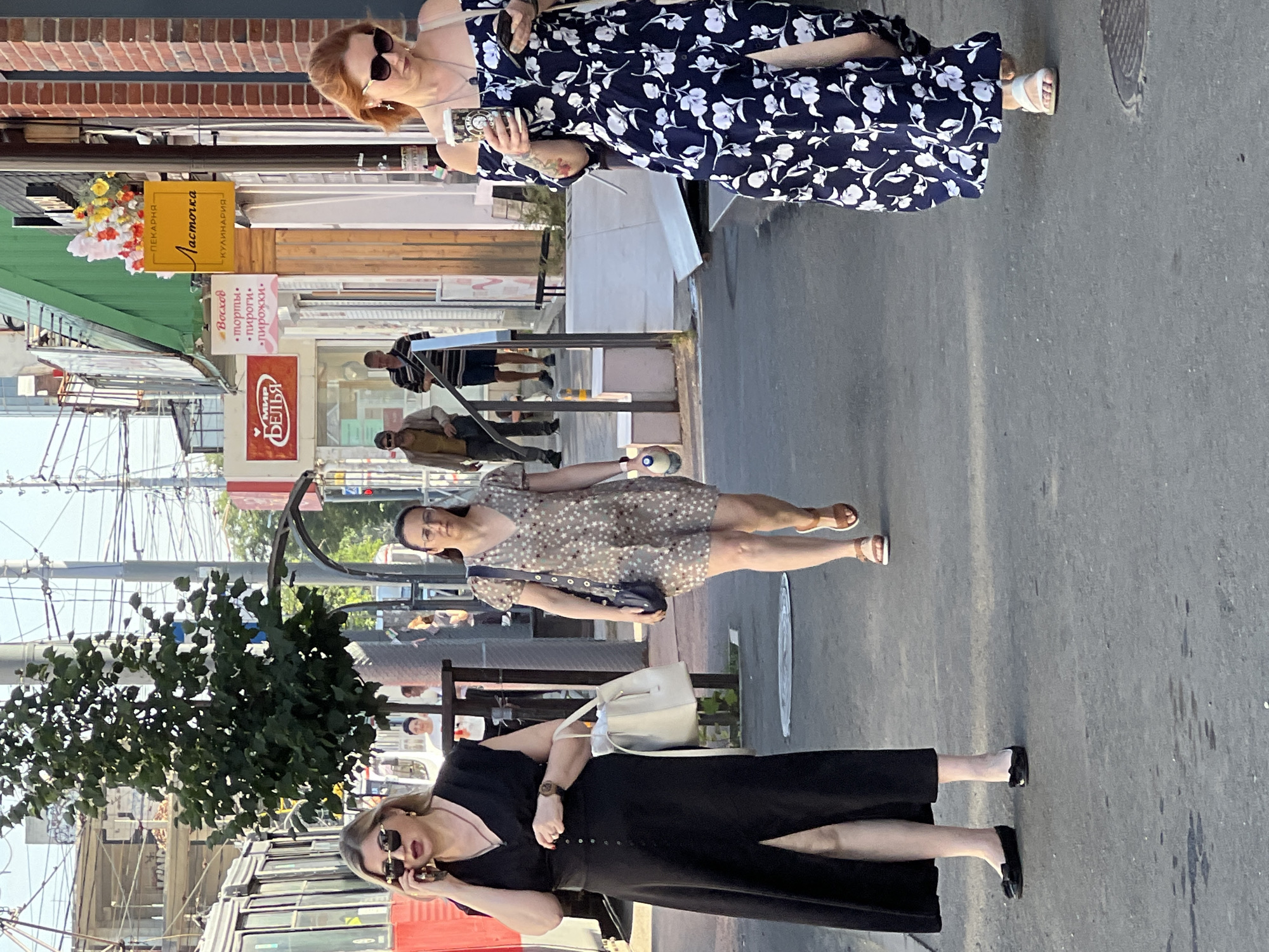 The image depicts a street scene in Krasnodar, Russia, likely near a transit stop, with several people walking. In the foreground, on the left, a woman wearing a black dress and sunglasses is talking on her phone while carrying a white bag. To her right, further down the sidewalk, a woman in a patterned dress is walking. On the far right, another woman in a floral dress is looking at her phone. In the background, several other individuals are visible, as is a tram or trolleybus on the left. The environment includes storefronts with signage, trees, and overhead power lines. The ground is paved with asphalt. The lighting suggests it is daytime, likely late afternoon or early evening due to the shadows.
