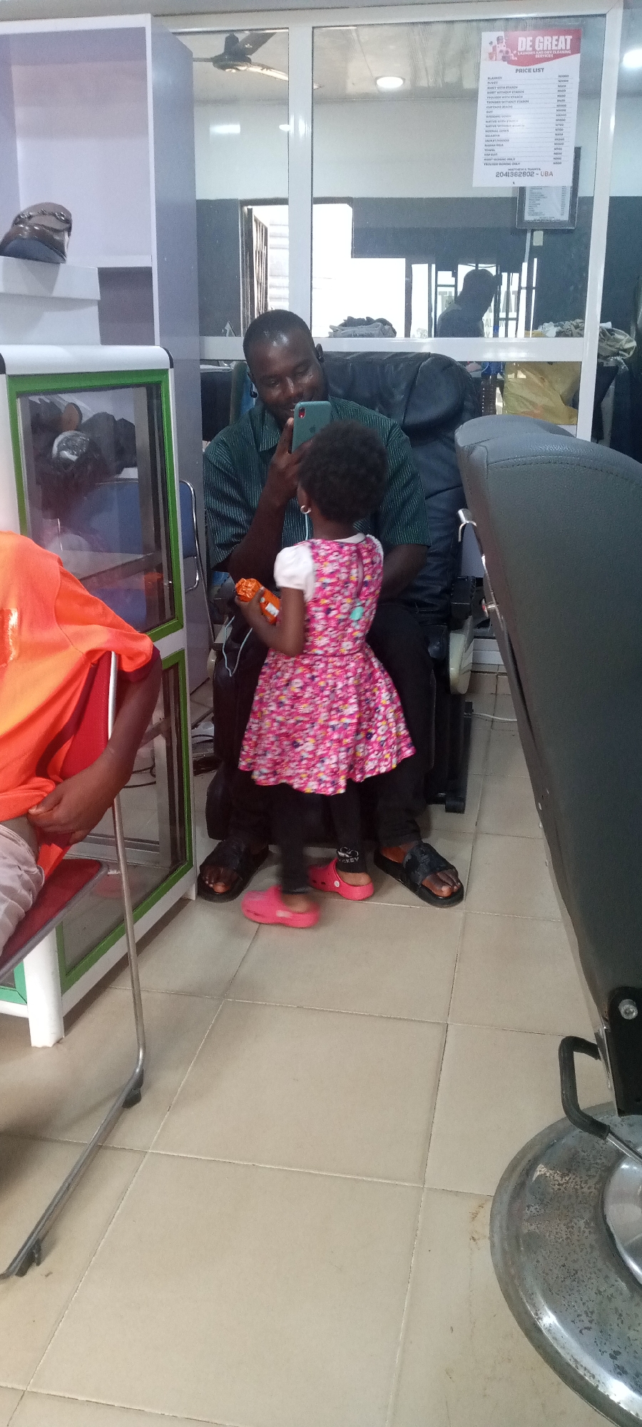 The image shows an adult man and a young girl inside what appears to be a business establishment in Jos, Nigeria. The man is seated, wearing a green patterned shirt, black pants, and sandals. He is holding a smartphone and appears to be taking a picture or video of the girl.
The girl is standing in front of the man, facing him. She is wearing a pink floral dress, black leggings, and pink Crocs. She is holding a small orange toy.
To the left of the man and girl is a glass display case containing objects that are difficult to discern clearly. Another person wearing an orange shirt is seated in a chair to the left of the display case. In the background, there is a mirror reflecting the scene, a price list hanging on the wall, and a partial view of the interior. A barber's chair is visible to the right. The floor is tiled.