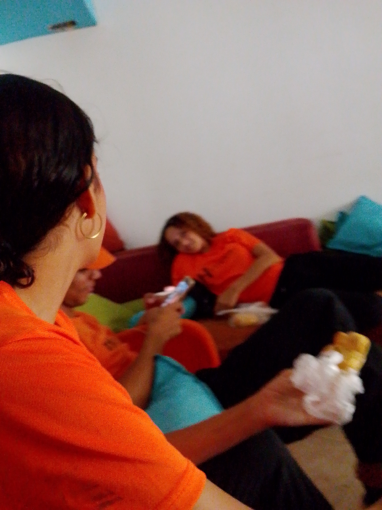 The scene depicts three individuals in an indoor setting, possibly a living room or common area. The location is Playa El Agua, Venezuela. The foreground features a person with dark hair and a gold hoop earring, wearing an orange t-shirt. They are holding an object wrapped in white material. Another person, also wearing an orange t-shirt, is partially visible and seems to be using a phone. A third person is lying on a maroon couch with turquoise pillows. The room has a white wall. The overall lighting suggests an indoor environment.