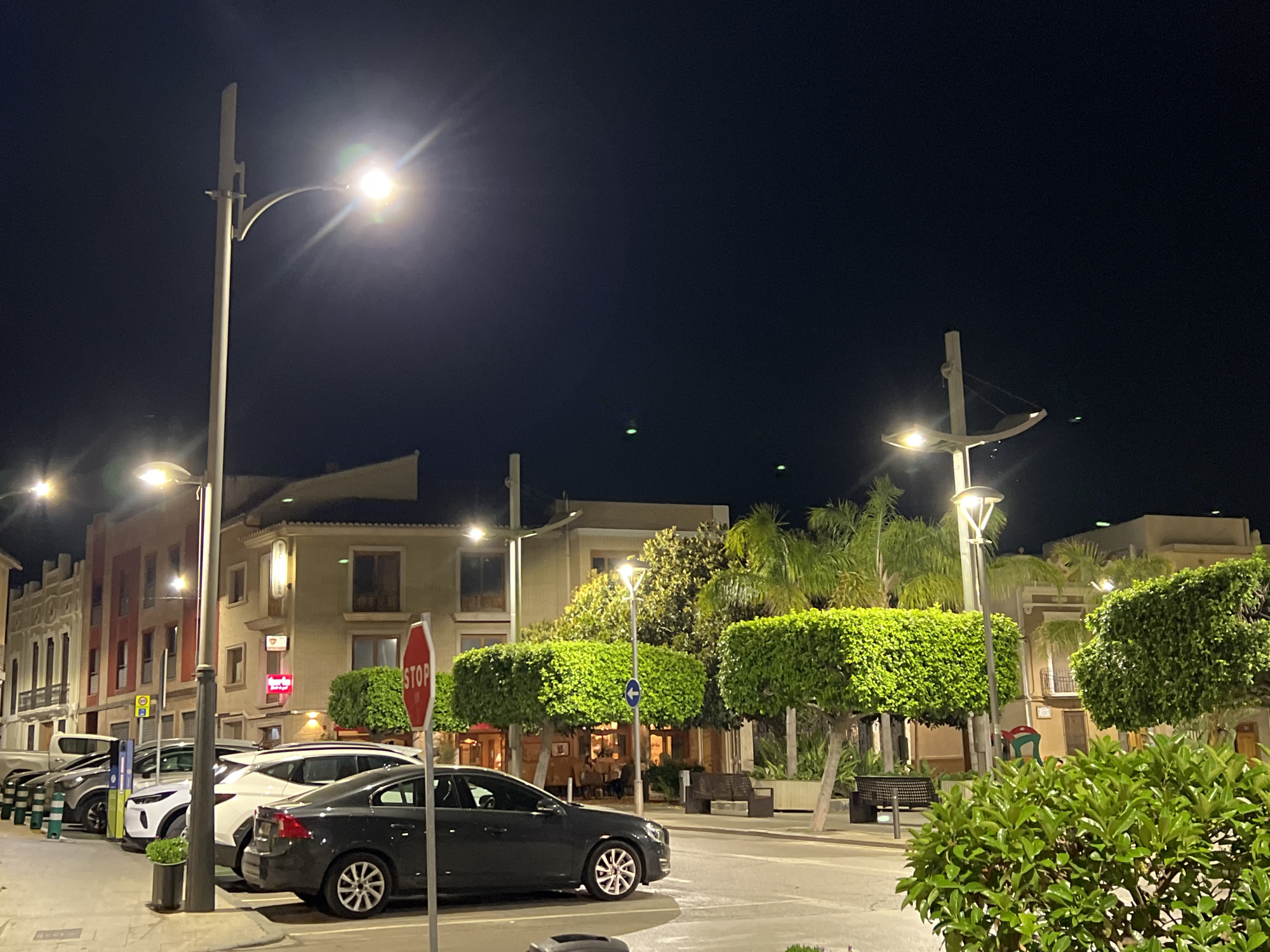 The scene is an urban street at night, likely in Picassent, Spain. Streetlights illuminate the area. Buildings line the street, with trees trimmed into box shapes planted alongside the road. Cars are parked along the curb. A stop sign is visible. There are benches along the sidewalk. No people are directly visible.