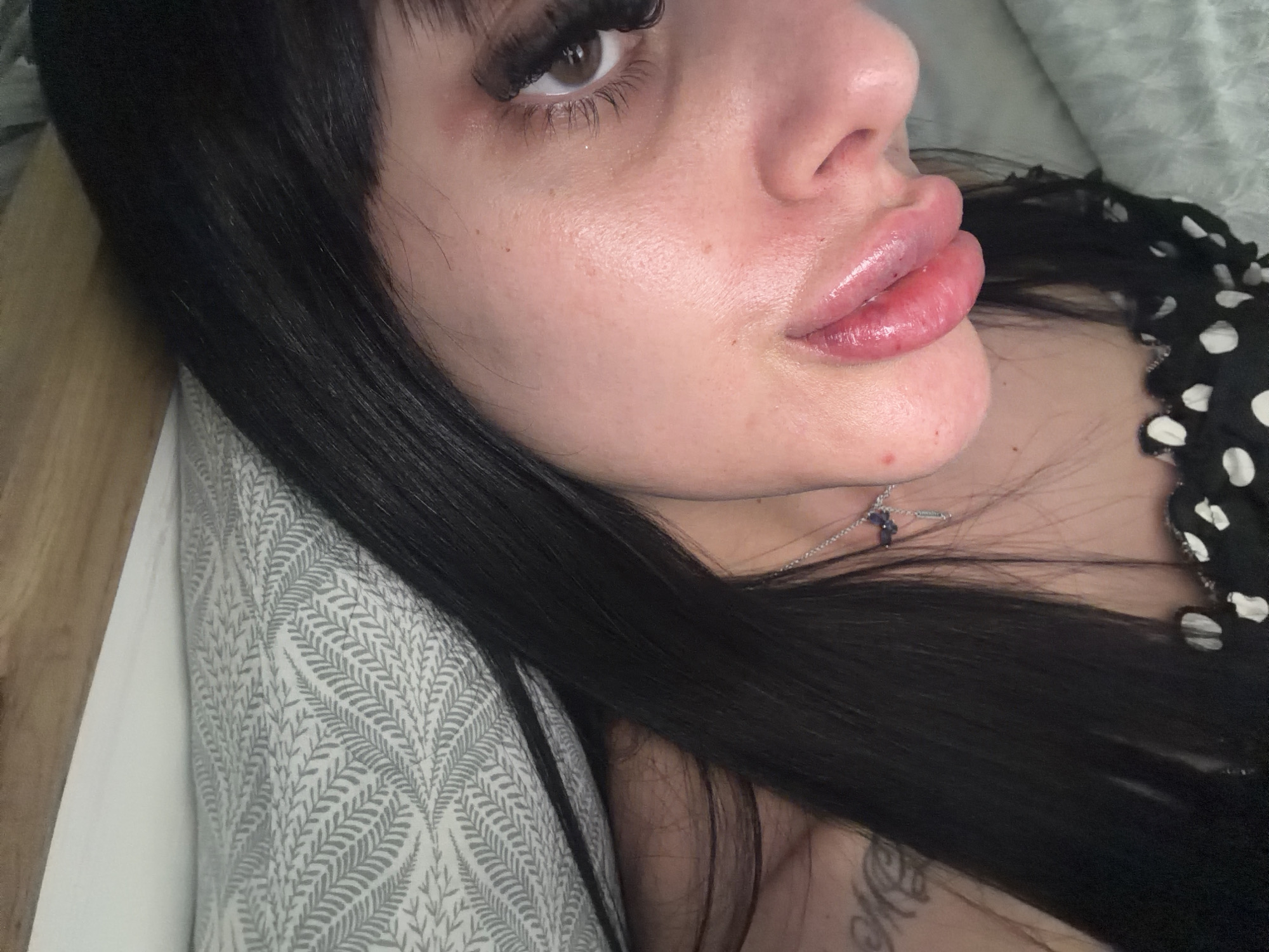 The image is a close-up, angled shot of a person's face, focusing on their lips, eye, and hair. The person has dark hair with bangs that fall across their forehead. They are wearing dramatic false eyelashes. Their lips appear to be augmented and are colored with a glossy, pinkish-red lipstick. A delicate silver necklace with a small, dark pendant is visible on their neck. The person is lying down, with their head resting on a pillow that has a subtle, grey leaf-like pattern. A portion of their black garment with white polka dots is visible on the right side of the frame. The background suggests an indoor setting, possibly a bedroom, with a soft, textured surface. The lighting is soft and diffused, highlighting the texture of the skin and the glossiness of the lips. There is no discernible indication of the city of Plovdiv, Bulgaria, or any specific location within it.