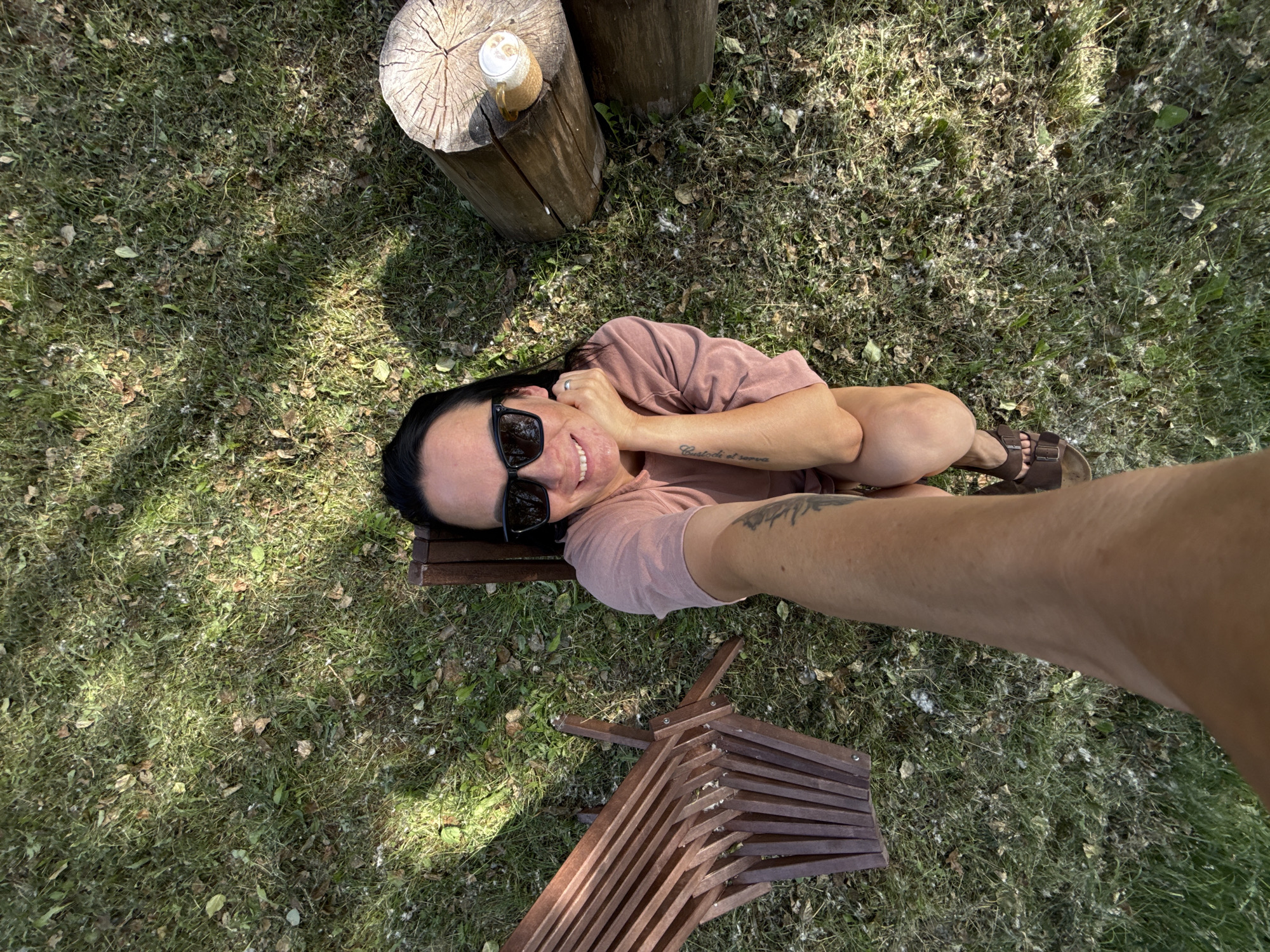 The image is a high-angle, selfie-style shot taken outdoors on a grassy area, possibly a park or backyard, in Chaikovsky, Russia. A woman with dark hair, wearing sunglasses and a pinkish-brown t-shirt, is the main subject. She is sitting on a wooden bench or chair. Her right hand is resting on her chin, and she is smiling at the camera. A tattoo is visible on her left arm. To her left is a folded wooden chair. To her right are two large, rough-hewn wooden logs, one of which has a glass of a frothy beverage on it. The ground is covered with grass, fallen leaves, and some white fluffy debris. Sunlight casts shadows on the ground.