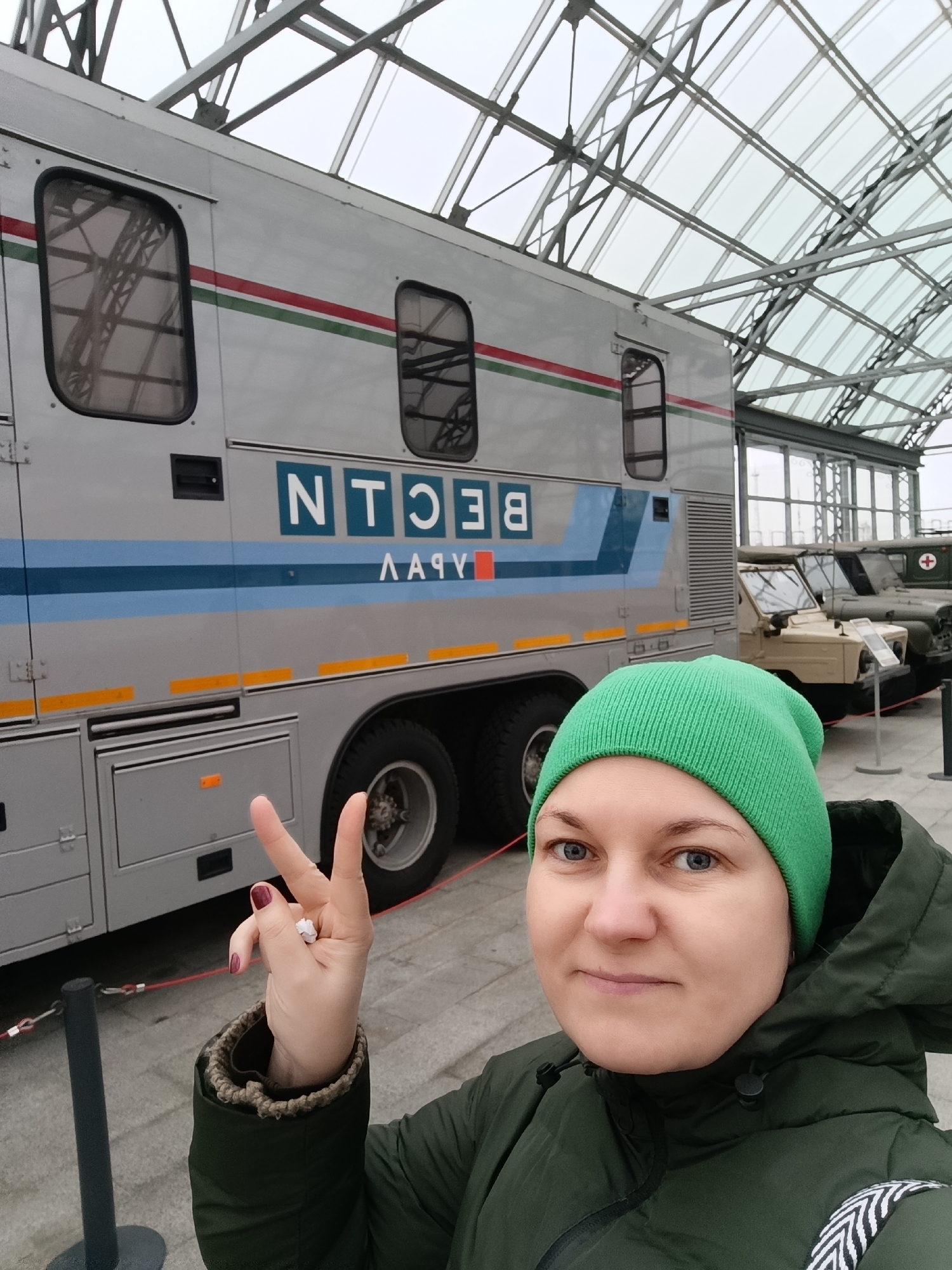 A woman takes a selfie inside what appears to be a museum or exhibition hall, likely the UMMC Museum of Military Equipment in Verkhnyaya Pyshma, Russia. She is wearing a green beanie and a dark green winter jacket, suggesting cool weather. Her expression is neutral as she holds up her left hand in a "peace" or "victory" sign, with a ring visible on her ring finger and dark nail polish.
Behind her is a large, silver, multi-axle vehicle, resembling a specialized truck or mobile command center. It features a horizontal blue stripe bordered by thin red and green lines along its side. Above the blue stripe, text is visible, appearing mirrored or reversed. When correctly oriented and interpreted as Cyrillic letters, the text reads "ВОТН" (VOTN). Below the blue stripe, similarly mirrored, the text "УРАВ" (URAV) is visible. Yellow rectangular markers are also present on the vehicle's lower section.
In the background, behind the large silver vehicle, several other military-style vehicles are displayed, including what looks like a light tan jeep and a green military ambulance with a red cross, indicating a collection of historical or military machinery. The entire display is housed under a distinctive transparent arched roof structure made of metal and glass, allowing for ample natural light. The floor is light-colored and paved.