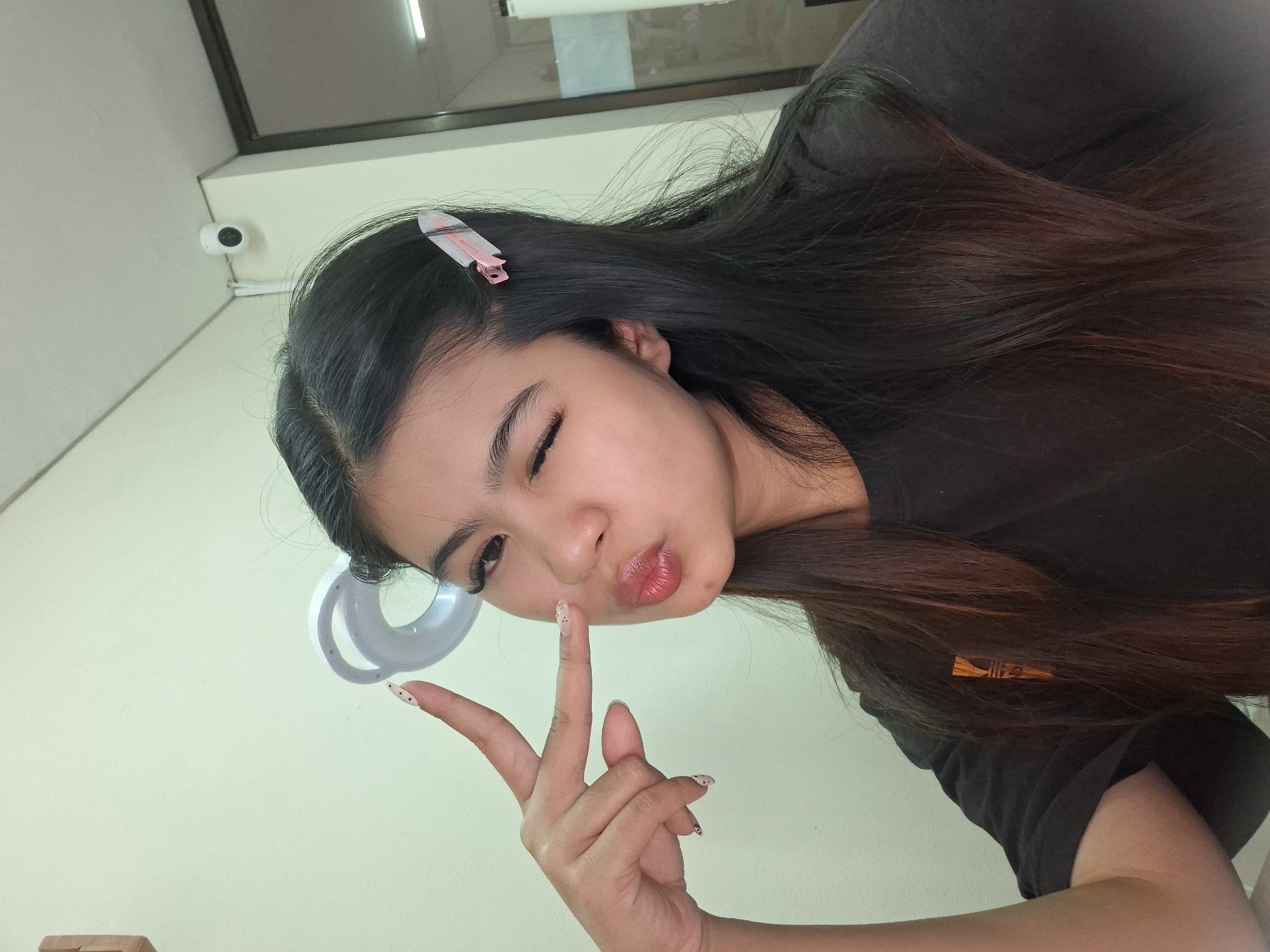 In Kuta, Indonesia, a young woman is captured in a selfie taken indoors. She is looking towards the camera, winking with her left eye, and puckering her lips. Her right hand is raised, with her index and middle fingers extended in a peace sign, positioned near her cheek. She has long, dark brown hair, some of which is pulled back with a pink hair clip. Her nails are painted with a light-colored polish and black polka dots. She is wearing a dark t-shirt. In the background, a ring light is visible, and a security camera is mounted on the wall. The wall behind her is a pale green color, and a door frame with glass is present on the right side of the image.