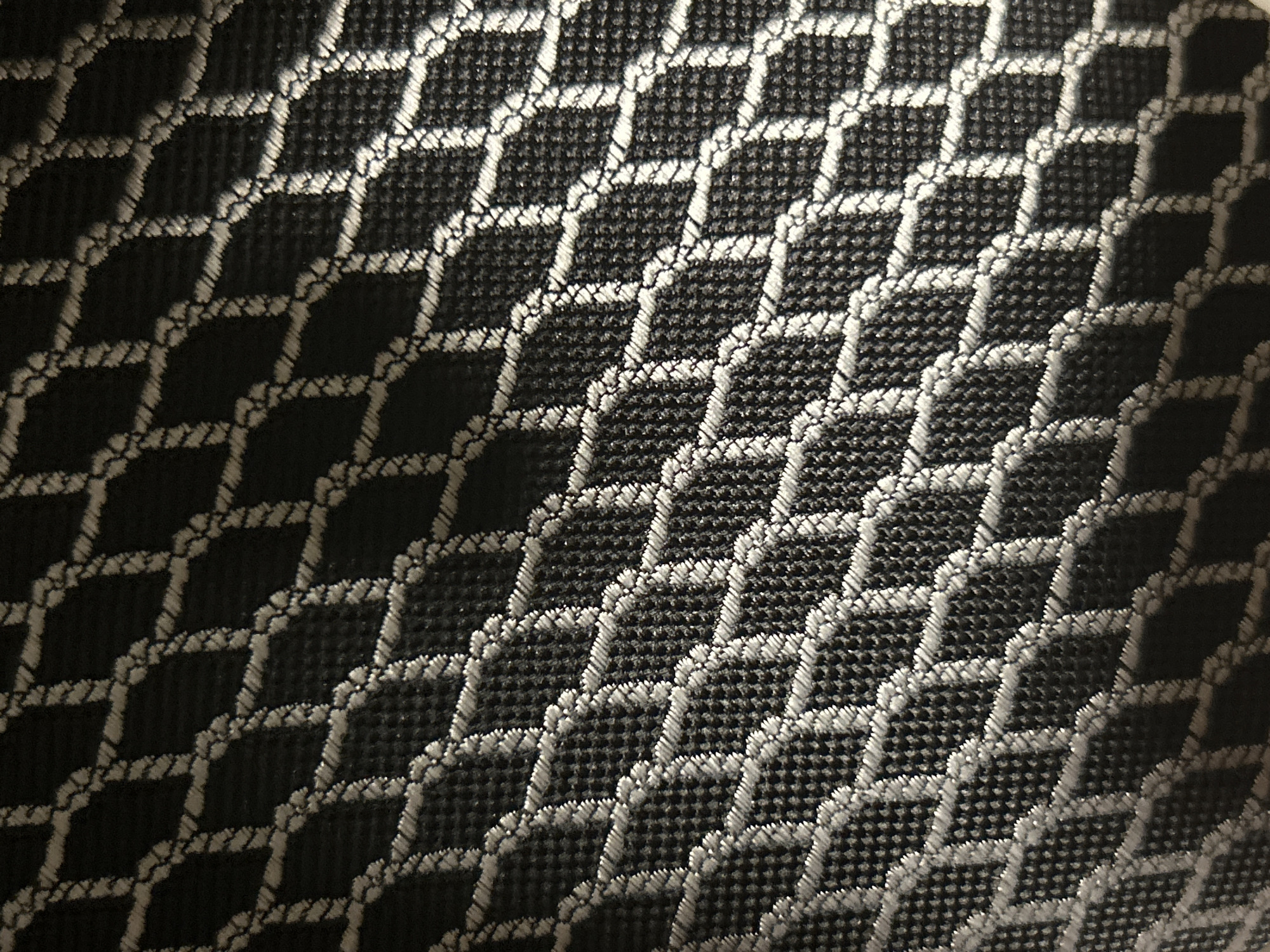 The image shows a close-up, angled view of a black fabric with a raised, white geometric pattern. The pattern consists of interconnected diamond shapes formed by a thicker, textured thread, creating a net-like appearance. The fabric itself appears to be a woven material with a visible, fine grid texture within the white outlines of the pattern. Shadows are cast by the raised pattern, adding depth and dimension to the surface. There are no people, specific locations, or actions depicted. This image does not provide any information about Moscow, Russia.