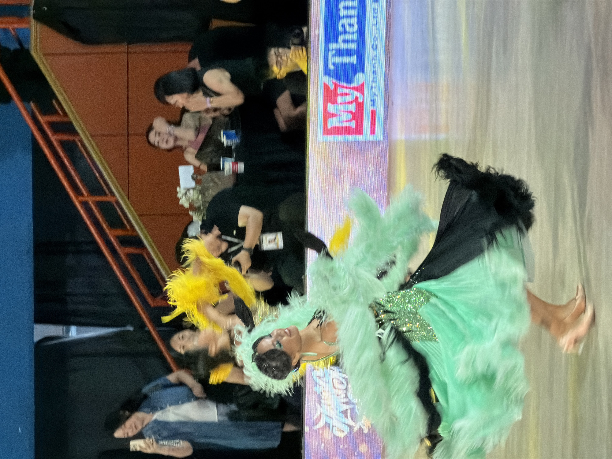 A female dancer in a shimmering green and black feathery costume is captured mid-motion, her body blurred by movement, on a polished floor that reflects light. She wears a sparkly green bodice and a voluminous green skirt with black accents, adorned with light green feathers that extend from her arms and neckline. Her face, partially obscured by her hair and costume, is turned to the side with a smile. In the background, several people are seated at tables, some watching the dancer. To the right, a large sign with red and blue lettering reads "My Thanh". The setting appears to be an indoor performance venue, potentially part of an event in Hà Nội, Vietnam, given the sponsor's name.