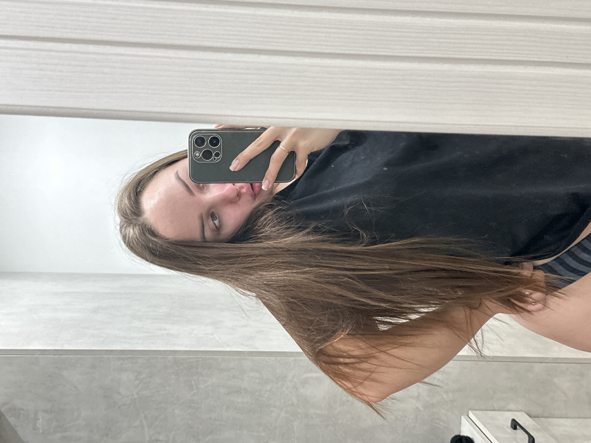 A woman is taking a selfie in a bathroom mirror. She holds a smartphone, partially obscuring her face. She has long, light brown hair. She is wearing a black t-shirt. The bathroom has white, tiled walls and a white cabinet with a black handle. The location is unspecified beyond being a bathroom setting. There is no indication of being in the city of Chaikovsky, Russia.