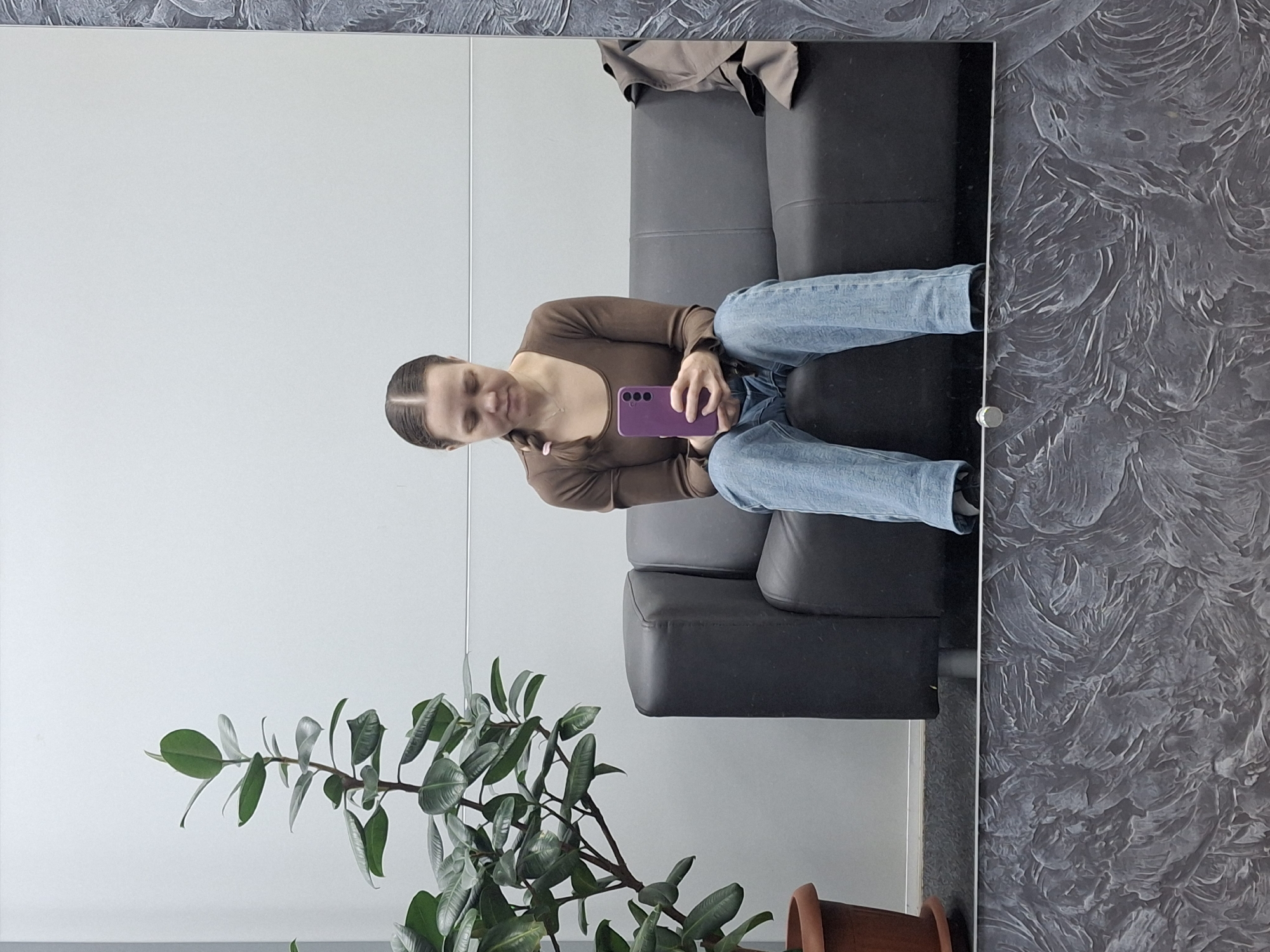 A mirror reflects a woman sitting on a black leather couch, holding a purple smartphone. She is wearing a brown long-sleeved shirt and blue jeans. To the left of the couch, in the foreground, is a potted plant with large, green leaves. Behind the couch, the wall is painted a solid white on the left and textured gray on the right. A tan jacket or coat is on the back of the couch. The reflection suggests the photo was taken indoors, likely in a waiting area or lounge. The location is Chaikovsky, Russia.