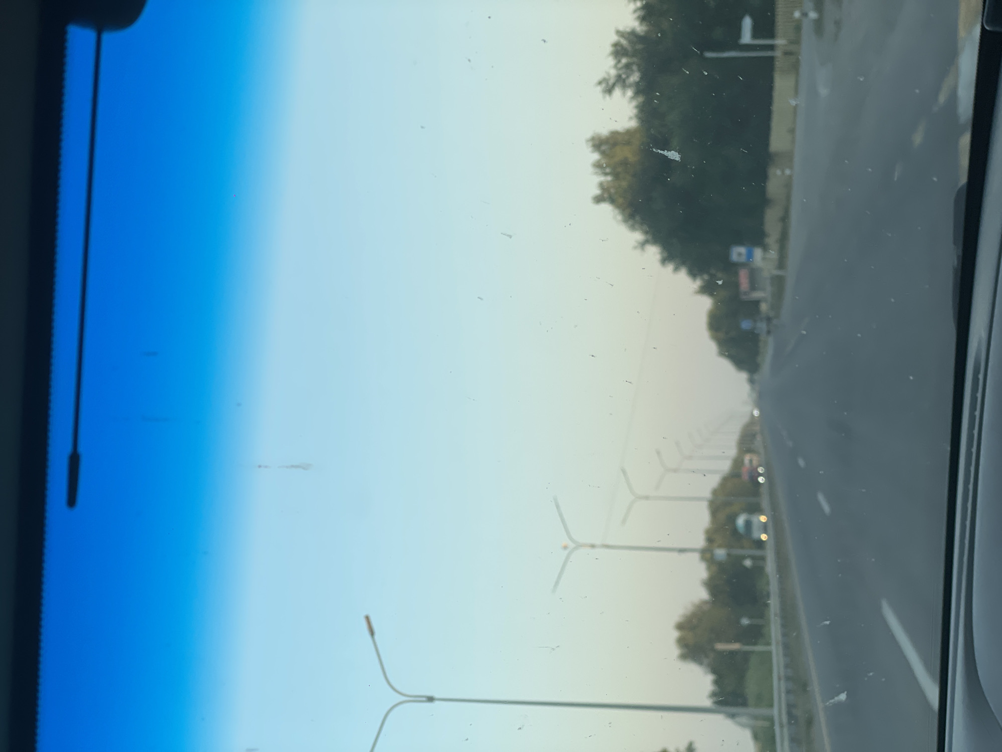 The image depicts a view from inside a vehicle traveling on a highway in the vicinity of Zhyemkum, Kazakhstan. The perspective is from the driver's seat, looking forward through the windshield. The highway is paved and has multiple lanes, with white and yellow lines marking the traffic lanes. Streetlights are visible along the sides of the road, and trees line the median and the shoulders. The sky transitions from a deep blue at the top to a lighter hue towards the horizon, suggesting it is either early morning or late afternoon. A few vehicles, including a white truck and some cars, are visible in the distance. The windshield has several smudges and specks, indicating it is not perfectly clean. The overall scene is consistent with travel on a rural or suburban highway.