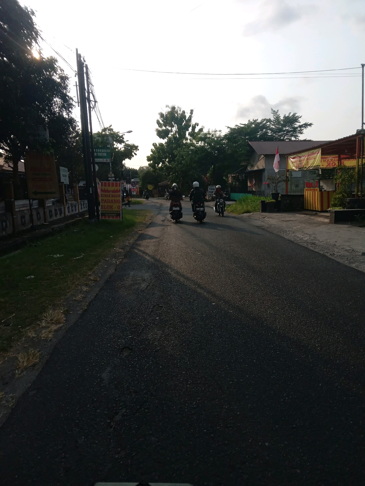 A photograph taken from a moving vehicle shows a street scene in Banguntapan, Indonesia, on a cloudy day with the sun setting, casting long shadows. The road is paved asphalt, with some patches and texture visible. On the left side of the road, there is a grass verge with some dry vegetation, and beyond that, a low concrete wall with a fence. Several utility poles with wires run along the left side of the road. Signs are visible, including a green sign with Indonesian text and a yellow sign with red text that appears to advertise "RUMAH MAKAN PADANG BLOK - O".
Several people on motorcycles are traveling along the road. Three motorcycles are clearly visible in the foreground and middle ground, each carrying a rider. The riders are wearing helmets, and one motorcycle has a passenger. The motorcycles are heading away from the viewer, towards the right side of the image.
On the right side of the road, there are buildings with tiled roofs. One building has a yellow awning with red and white striped elements and appears to be a business. A flag, possibly the Indonesian flag, is visible on one of the buildings. Trees are present on both sides of the road, with some lush green foliage and some dry branches. The overall impression is of a typical street in a semi-urban or suburban area of Indonesia during the late afternoon.