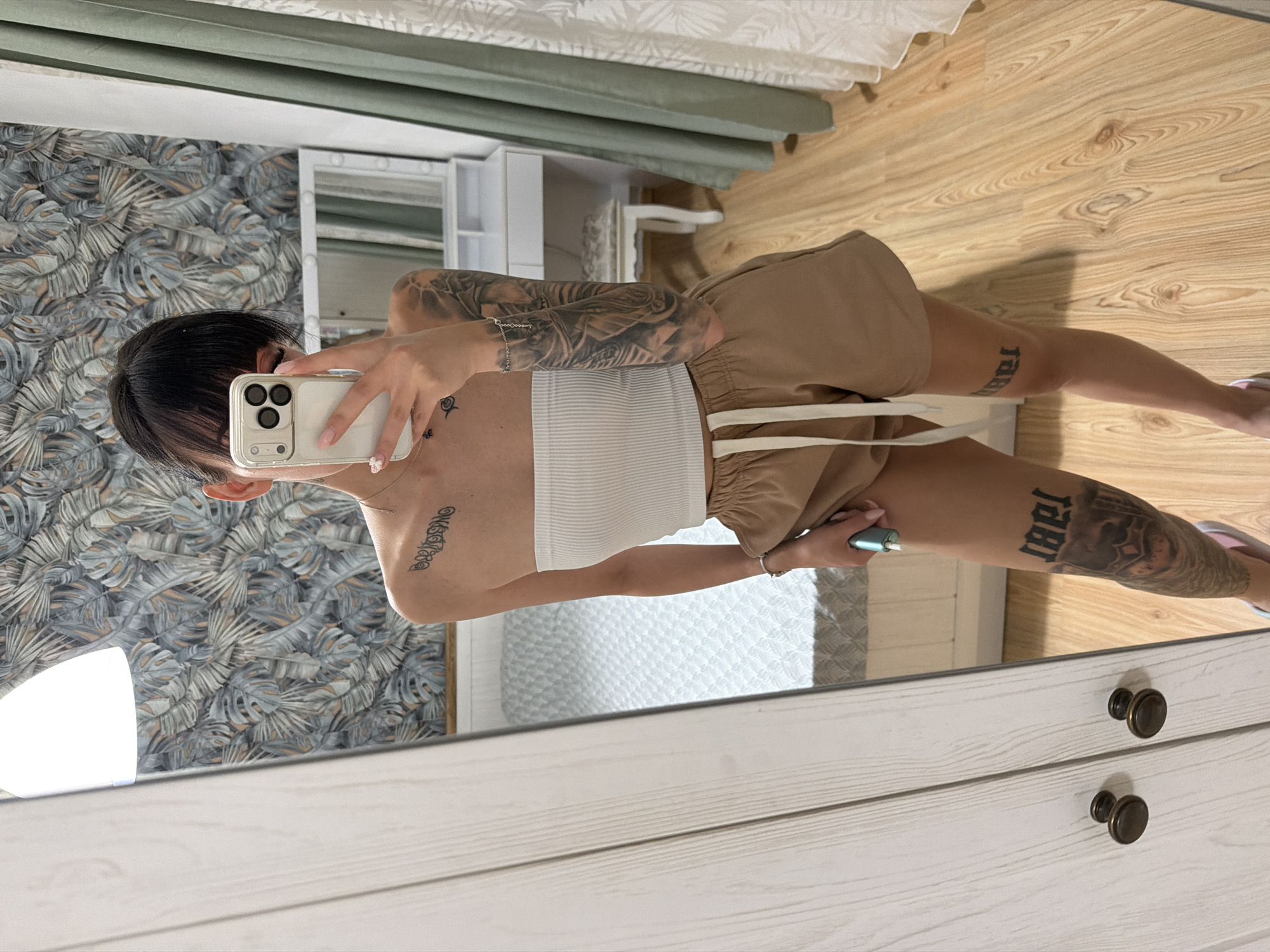 A young woman with dark hair and bangs is taking a mirror selfie in a bedroom. She is wearing a white strapless top and tan shorts. She has extensive tattoos on her arms and legs. The room has patterned wallpaper, a bed, a vanity with a mirror, and light-colored wooden floors. The image appears to be taken indoors, possibly in an apartment. There is no information in the image to suggest it is located in Plovdiv, Bulgaria.