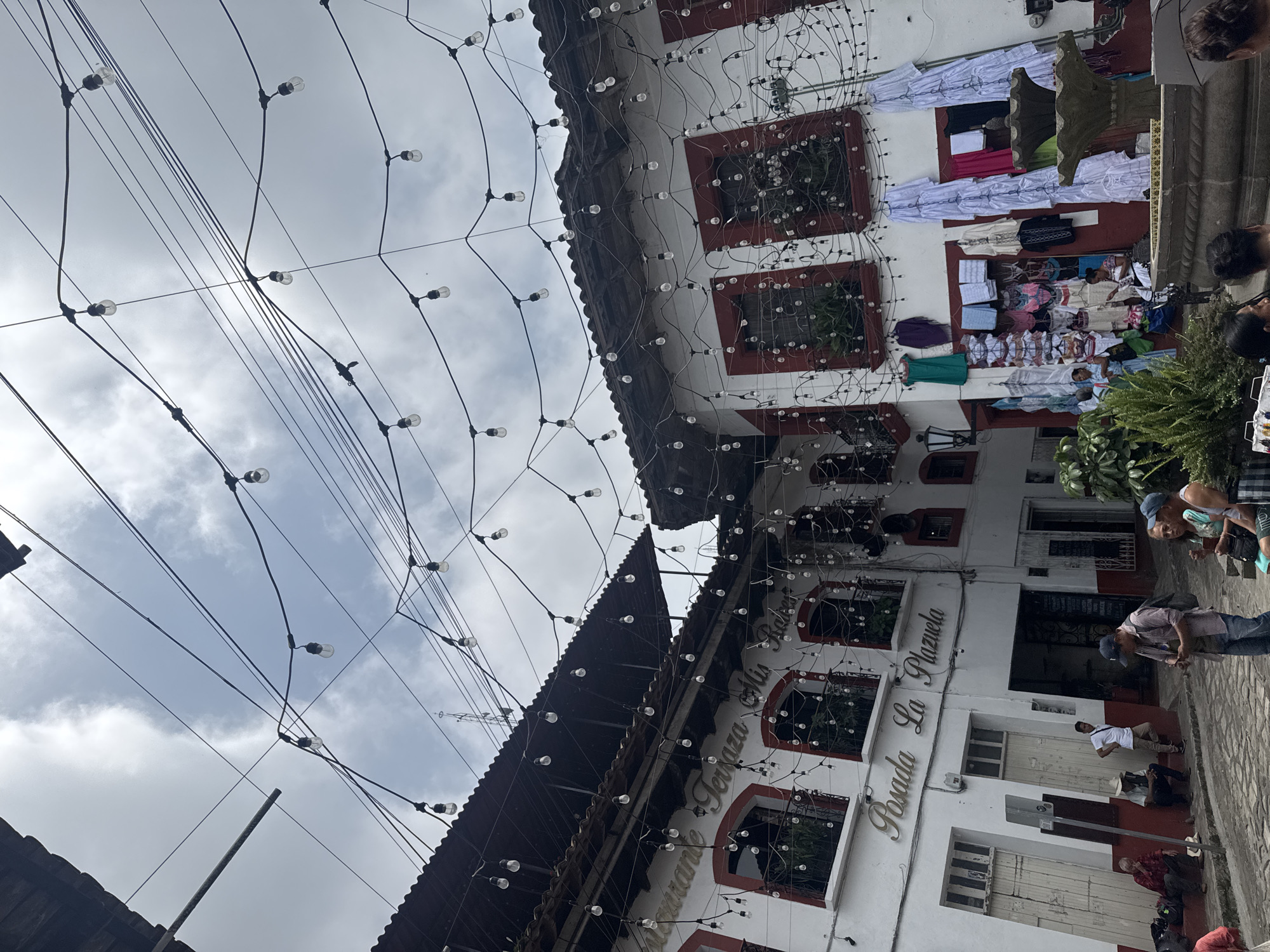 The image captures a street scene in Cuetzalan, Mexico. The view is directed upwards, showcasing a network of light bulbs strung across the street. On either side, buildings are visible. One building has a sign reading "Restaurante Terraza Mis Rios Posada La Plazuela." Clothes are hanging on another building's balcony. People are present on the street, some appearing to be vendors and shoppers. The sky is overcast.
