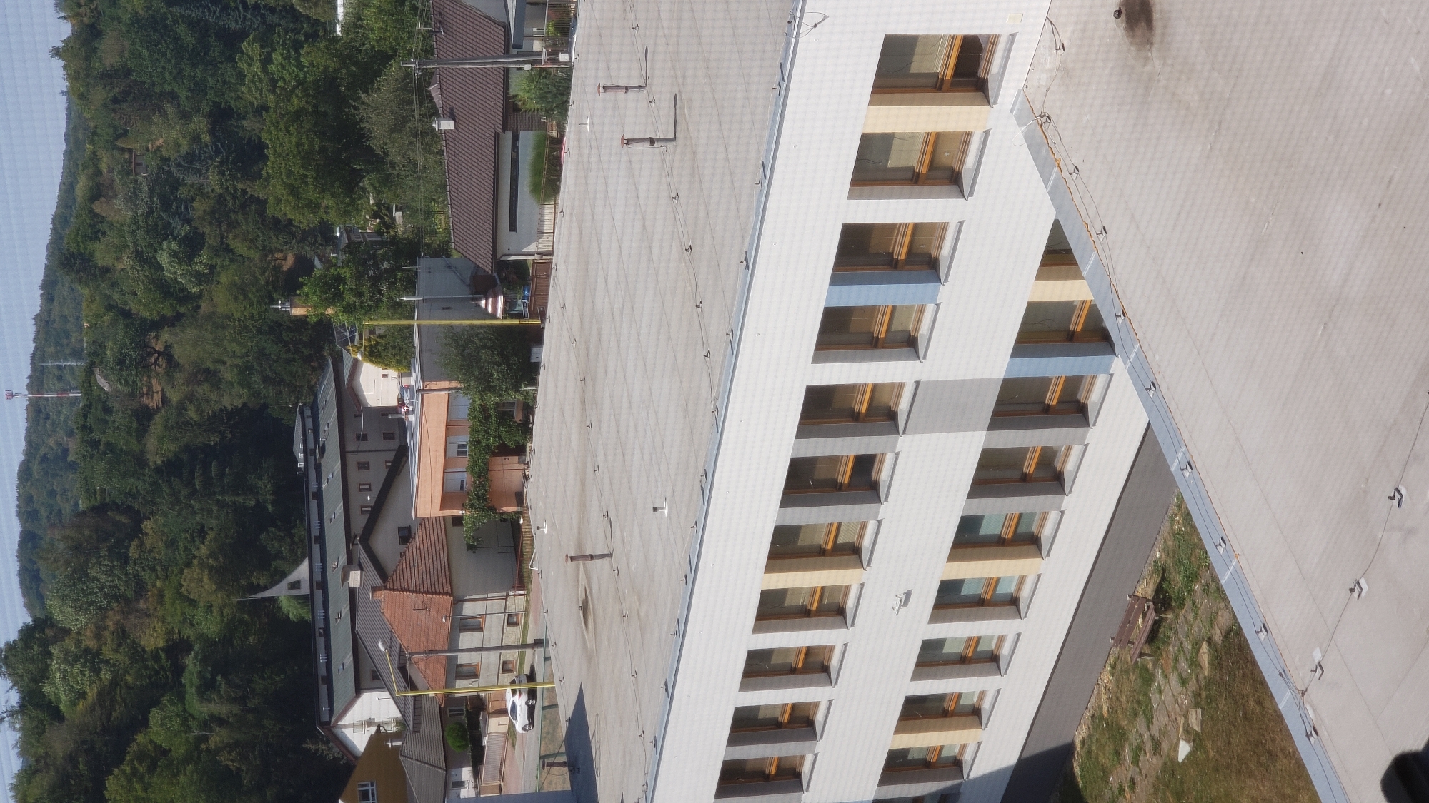 The image shows an aerial view of a building with a flat roof and multiple rows of windows. The building appears to be a school or administrative facility, with a simple, modern design. The windows are large and rectangular, with wooden frames. Some of the window panes are reflecting the surrounding environment. The roof is mostly flat, with a few small structures and vents. There is a small patch of grass and a wooden bench in front of the building. In the background, there are several other buildings and trees, as well as a hill covered in vegetation. The sky is clear and blue. The overall impression is one of a well-maintained urban environment.