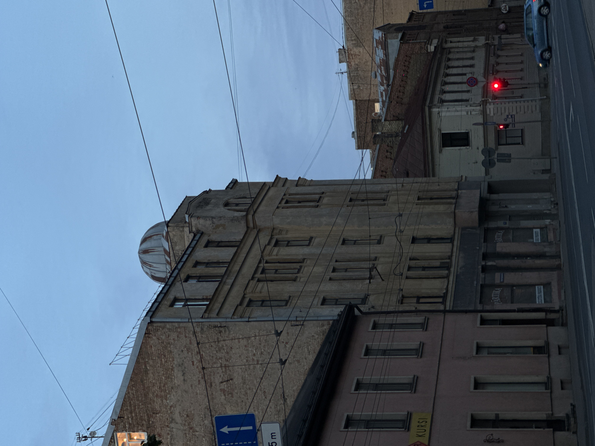 This media file captures a street scene in Riga, Latvia, with a focus on the architecture of the buildings. The image is oriented vertically, suggesting a perspective looking upwards or sideways. The sky is overcast with a muted blue hue, indicating either early morning or late evening.
The dominant subjects are several multi-story buildings. The central building, made of brick, features a distinctive dome-like structure at its apex, partially covered with a striped pattern. It has numerous windows and architectural embellishments. To the right, a lighter-colored building with a more modern facade is visible, featuring multiple rows of evenly spaced rectangular windows. Adjacent to this, another building with a darker, reddish-brown brick exterior is seen.
A street runs along the right side of the image, with a car driving by and a traffic light displaying a red signal. Overhead, a network of power lines crisscrosses the sky, a common sight in urban environments.
Notable details include a blue traffic sign with a white arrow pointing left, and a smaller sign below it with the text "5 m". On the facade of the lighter-colored building, signs with the text "PARTIKA" are visible, likely indicating businesses or establishments within. Another yellow sign with the text "KURSI" is also present. The overall mood is somewhat subdued, consistent with the twilight lighting conditions. No people are clearly visible in the frame.
