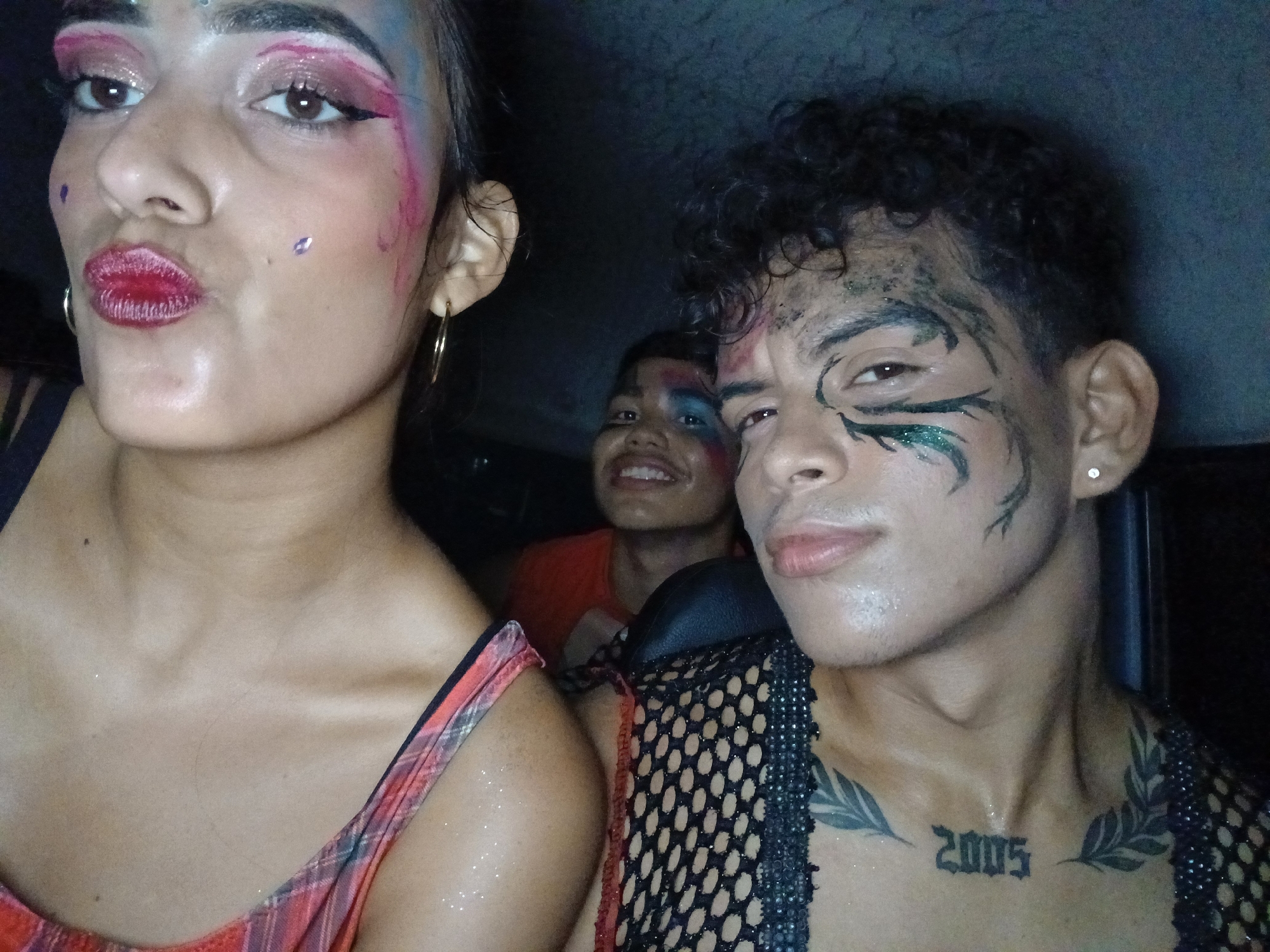 Three individuals are visible in this indoor shot. The first individual, a person with reddish-pink eye makeup, is positioned on the left, with a pouting expression. This individual is wearing a red and black checkered top and gold hoop earrings. The second individual, on the right, displays dark curly hair and green makeup around their eyes. A black, open-net shirt is worn, and a tattoo with the number "2005" can be observed on their chest. The third individual is situated in the background between the other two individuals and has blue face paint. All three individuals appear to be in a car. The location is unknown, but the request specified Playa El Agua, Venezuela. However, there is no indication of the city in the image.